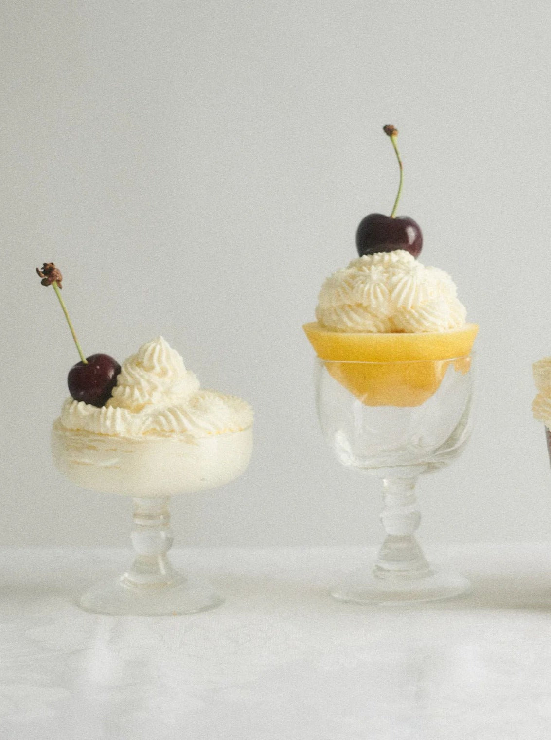 Two elegant Akua Objects Alban Wine Glass - Set of Four dessert cups filled with whipped cream and vanilla custard, each topped with fresh cherries, on a light background.