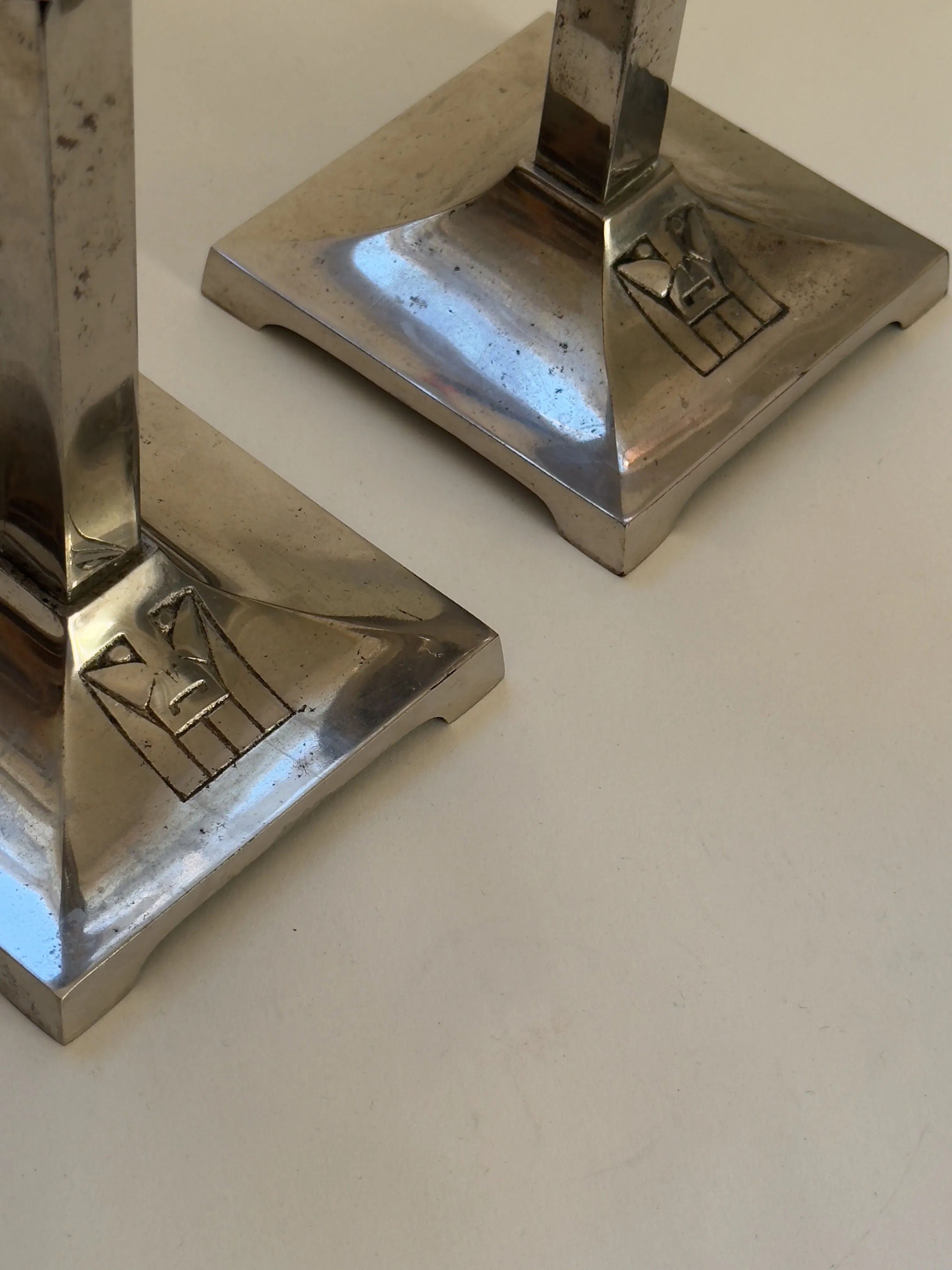 Galerie Storm's Pair of Art Deco Candlesticks from the 20s/30s feature two metallic bases with square designs, evoking an early modernist aesthetic. The engraved symbols resemble abstract geometric icons on a reflective surface with tarnished areas.