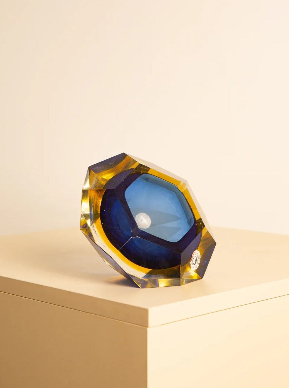 A faceted, multi-colored Large vide-poches blue "Diamond" by Flavio Poli 60's from Treaptyque with shades of blue, yellow, and green sits on a beige surface. The handcrafted piece has an asymmetrical geometric shape, and the light refracts through it, creating a dynamic visual effect.