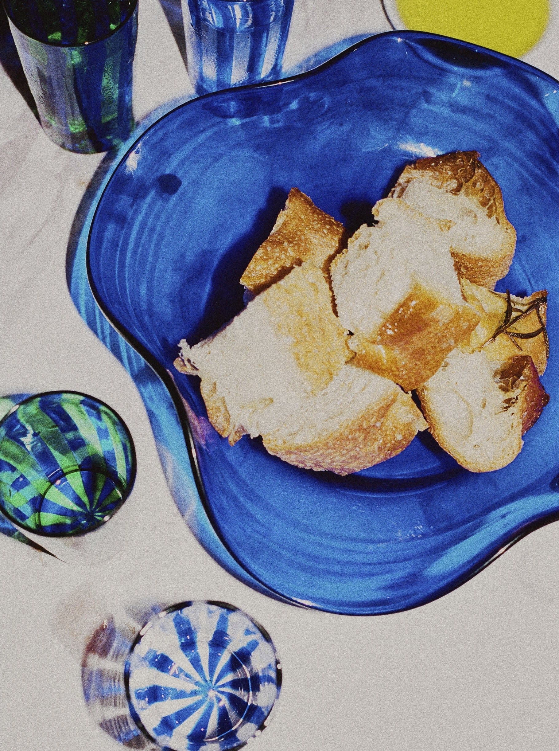 A scalloped blue glass dish filled with pieces of bread sits on a table. The table also features several glasses from the Michele Collection with blue and green swirling patterns and a white Akua Objects Michele Bowl Large with a yellow liquid, possibly olive oil. This exquisite Venetian glassware scene is well-lit with natural light.