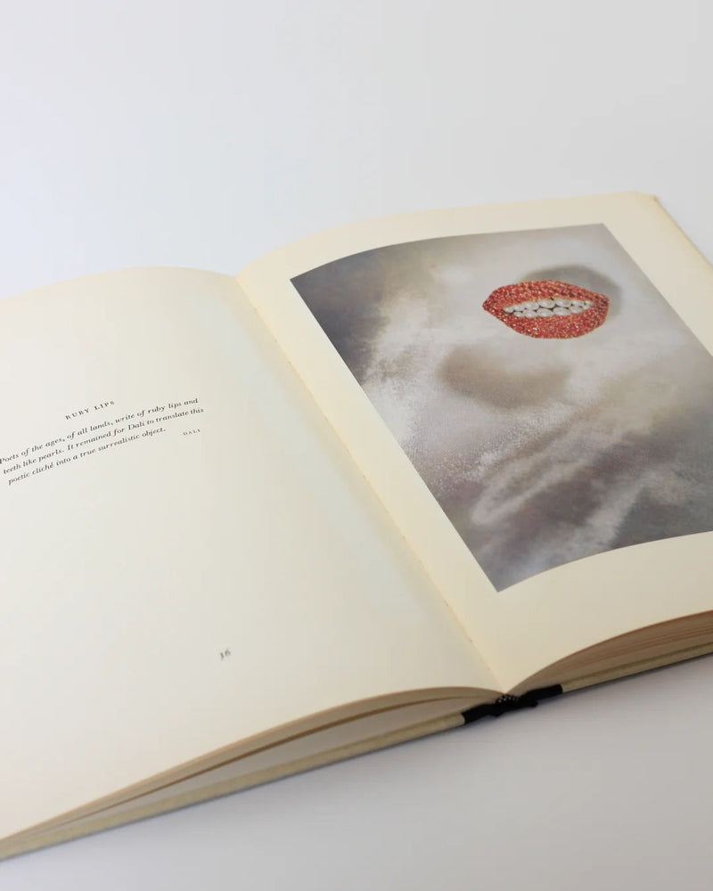 An open book on a light surface displays the left page with text under the header "REST UP," while the right showcases an abstract image of lips against a cloudy background, reminiscent of Dalí's surrealist style, from "Dalí: A Study of his Art-in-Jewels" by Boga Avante Shop.