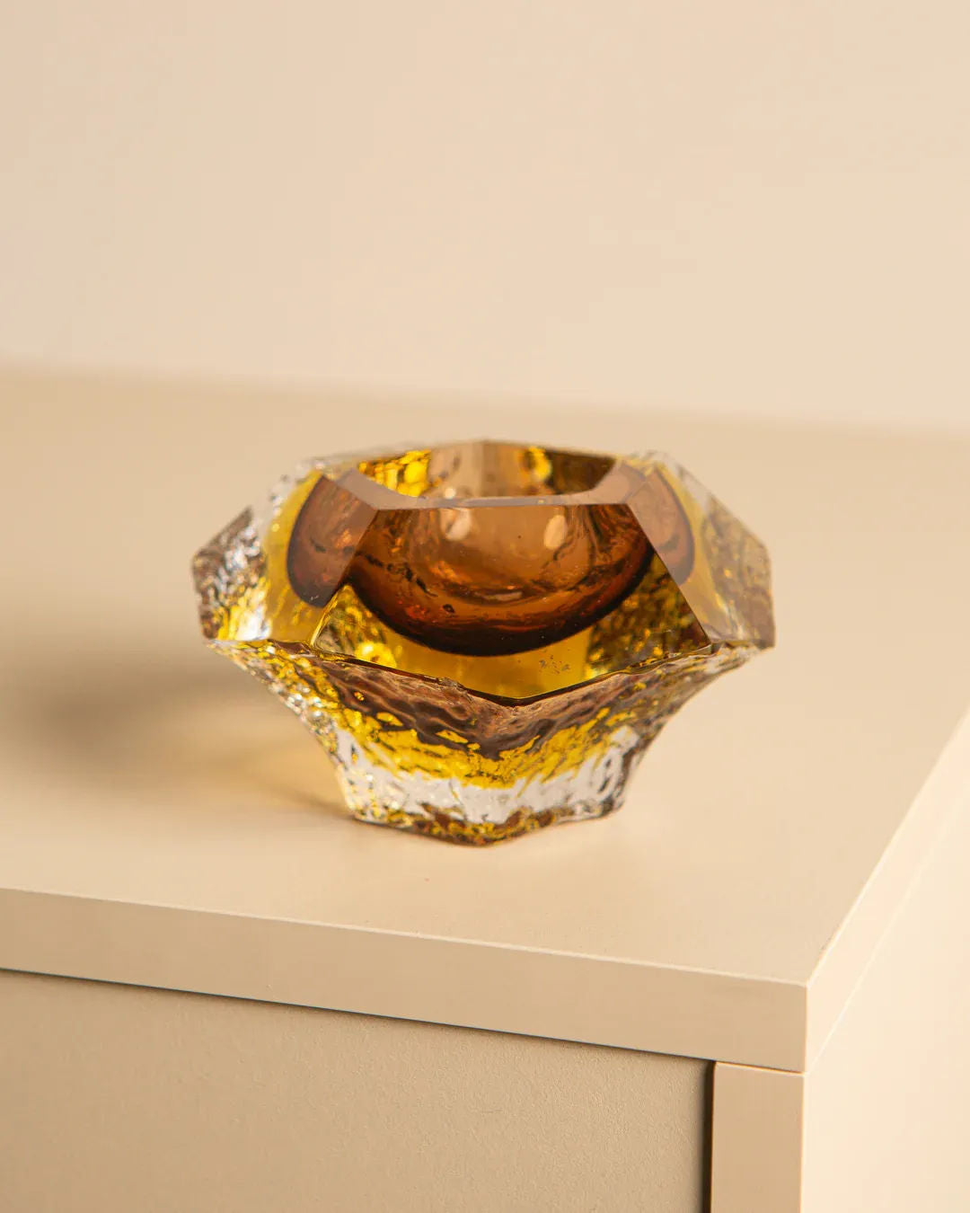 A faceted glass bowl, resembling Treaptyque's 'Diamant' Ashtray Murano by Flavio Poli for Seguso 60’s, sits on a light tabletop. Its warm amber hue and geometric angles capture the light, creating a shimmer that blends elegance with functionality.