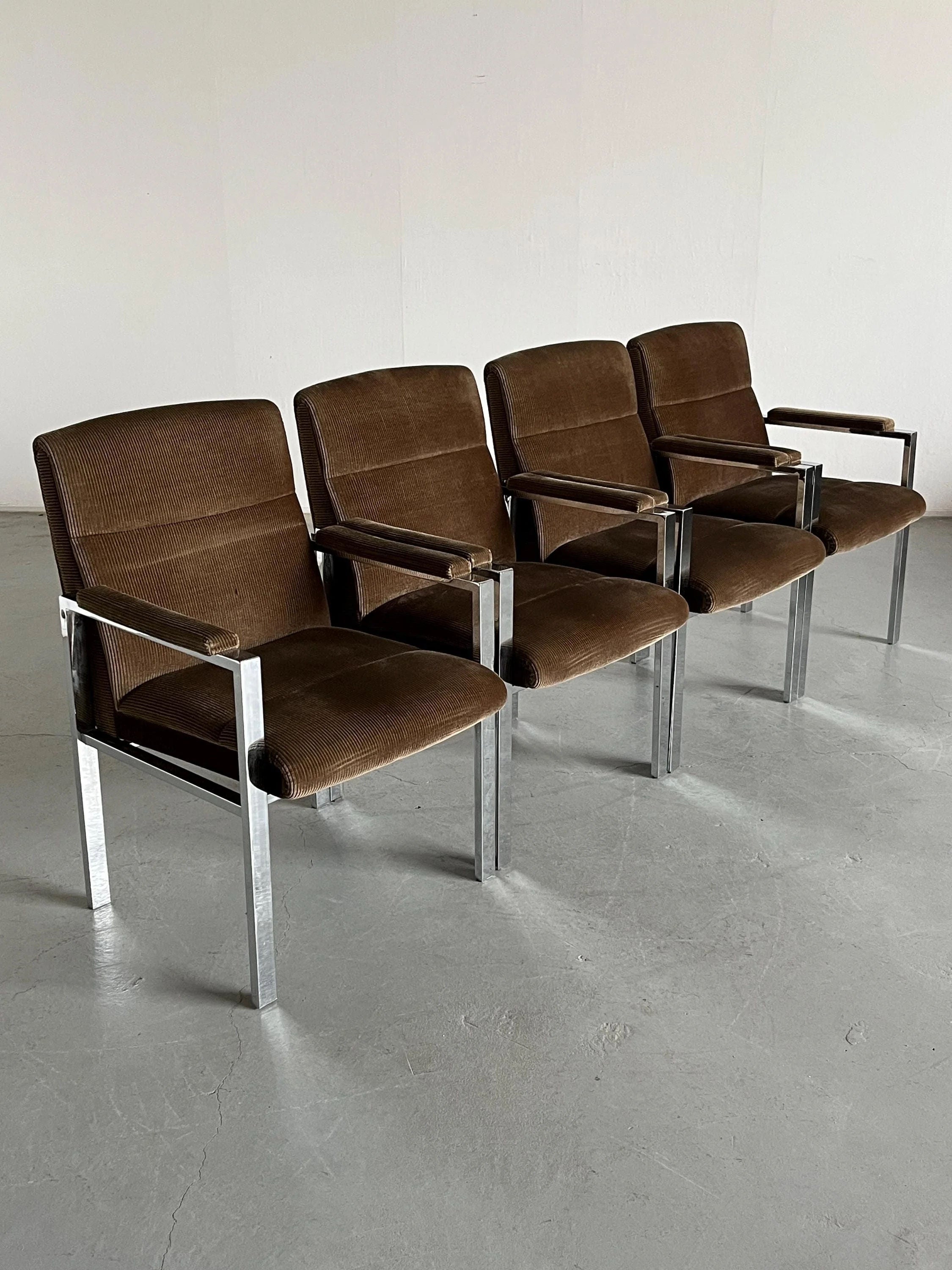 Four Mid-Century Armchairs by Cherry Cargo, featuring brown corduroy upholstery and chromed steel frames with armrests, are lined up on a gray floor against a plain wall. Their simple design embodies a minimalist aesthetic reminiscent of 1970s Vintage Mid-century modern style.