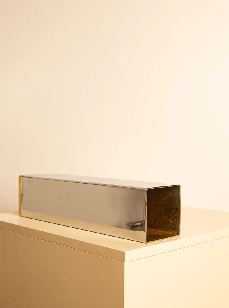 An elongated, rectangular metal box with an open end, likely made of mirrored stainless steel, lies horizontally on a light beige surface against a matching beige background. Its simple, minimalistic design evokes a touch of 70s elegance similar to the Large “Colonne” Vase 70's by Treaptyque.
