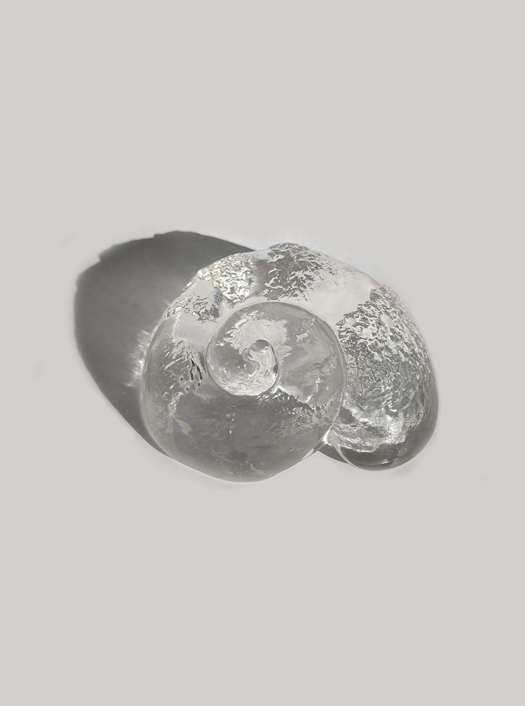 The Glass Shell Vide-Poches by Les Objoies is a clear glass piece shaped like a fluid swirl. Its heavy textured surface reflects light beautifully, casting a soft shadow against a light gray background.