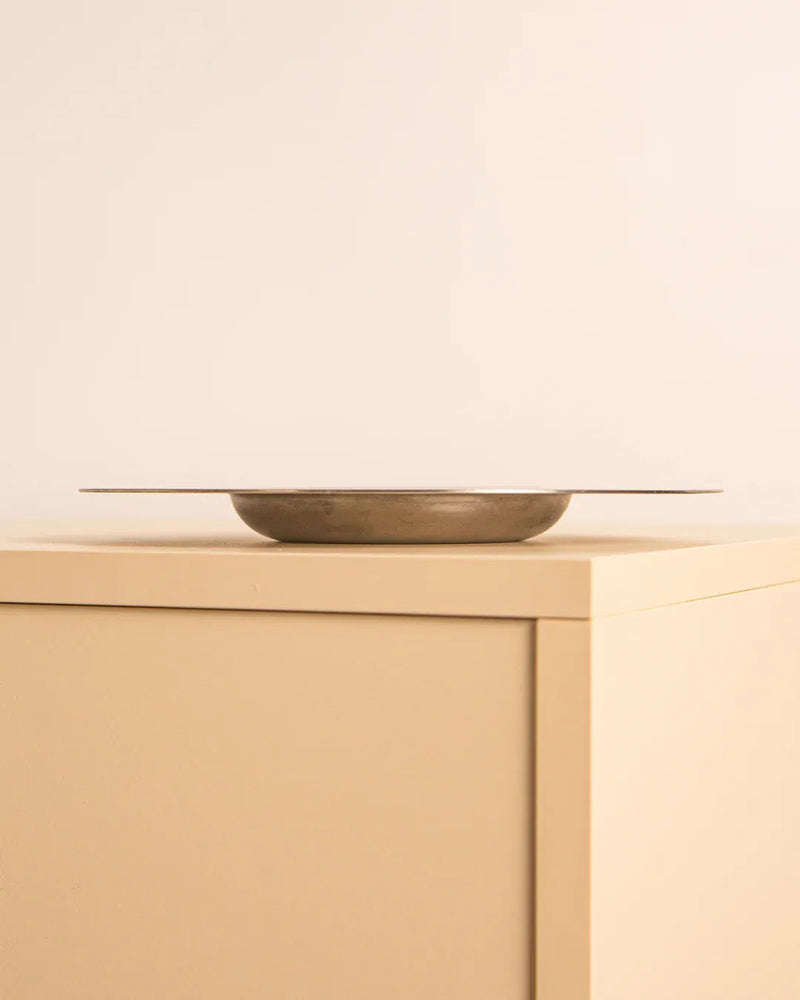 The Italian Circular Ashtray by Velca 80's from Treaptyque, with its minimalist, shallow design and metallic finish, conjures a vintage feel as it rests on a beige, square piece of furniture against a neutral, light-colored wall.