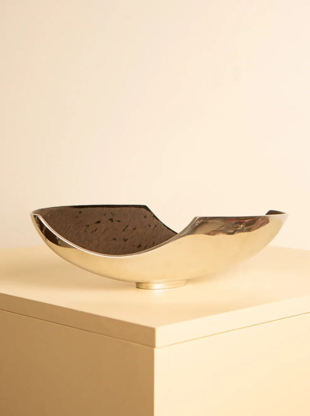 A metallic, asymmetrical bowl with a polished, reflective surface rests on a minimalist, light-colored pedestal against a plain beige background. This elegant piece boasts curved edges and an irregular shape reminiscent of an 80's fruit bowl design. The Treaptyque 80's Metal Fruit Bowl adds a touch of timeless sophistication to any setting.