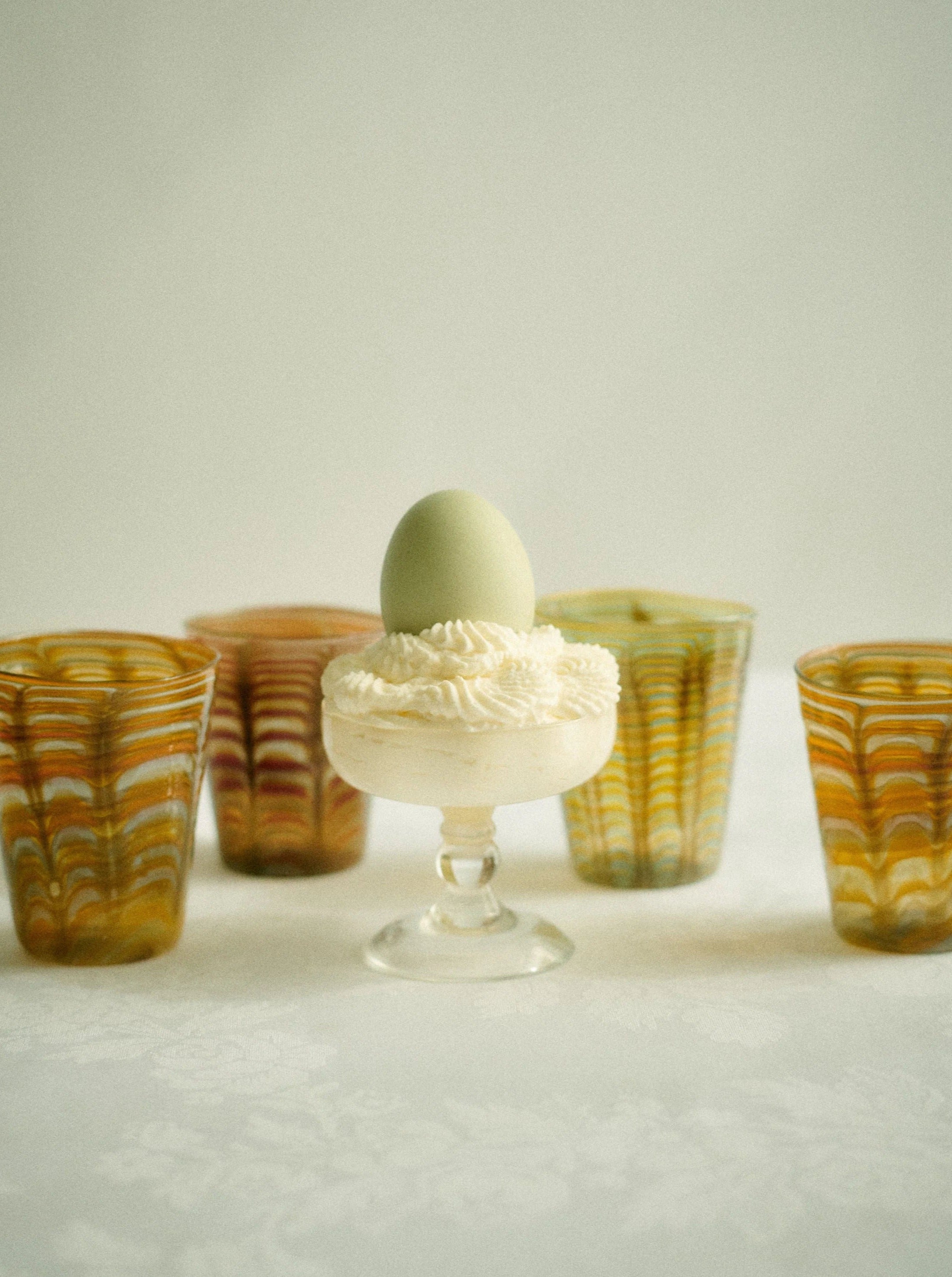 A soft-boiled egg atop a small Alban Champagne Glass egg cup from Akua Objects, surrounded by four colorful Bohemian Glassware cups, set against a light, textured background.