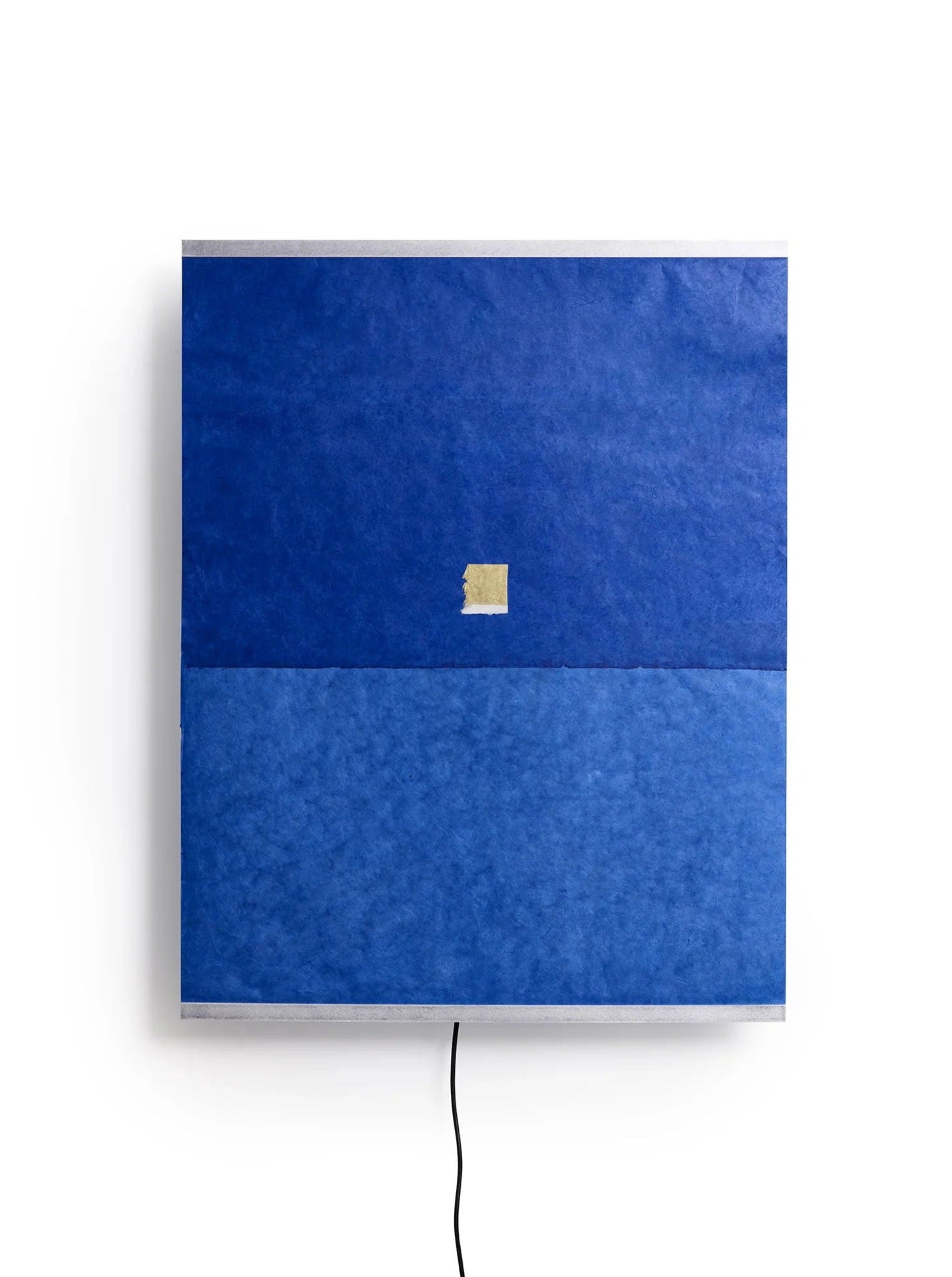 The Panel Wall Lamp – Montenegro by TEGET is a square piece that echoes the design aesthetic of Ana Kraš. It displays two shades of blue, with a lighter shade on the bottom and a darker one on top. Near the center, a small beige square is affixed, while an elegant black cord hangs below.