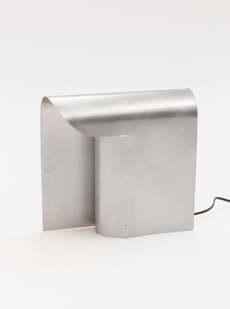 A sleek, modern metallic light fixture with a minimalist design, featuring a curved sheet of brushed stainless steel standing upright. The elegant and contemporary appearance is complemented by an E27 socket for versatile bulb options, with a black electrical cord partially visible on the right side. This is the Arc Table Lamp by Paul Coenen.