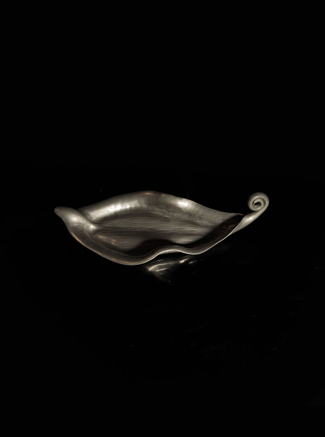 Set against a black background, the Handmade Leaf Vide Poche by Les Objoies features a decorative pewter design with curved edges and an elegant spiral handle. Its smooth surface subtly reflects light, evoking the style of an Art Nouveau vide-poche.