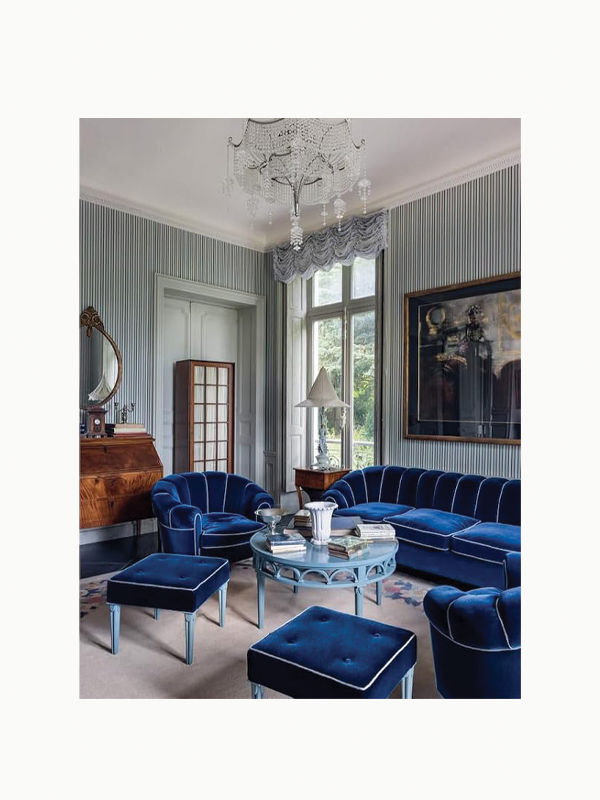 An elegant living room, reminiscent of an art deco mansion, features blue velvet furniture including a sofa, chairs, and ottomans. A glass coffee table with a white vase sits in the center, accompanied by Karl Lagerfeld: A Life in Houses from Maison Plage. The room has a large window, striped wallpaper, chandelier, and framed painting on the wall.