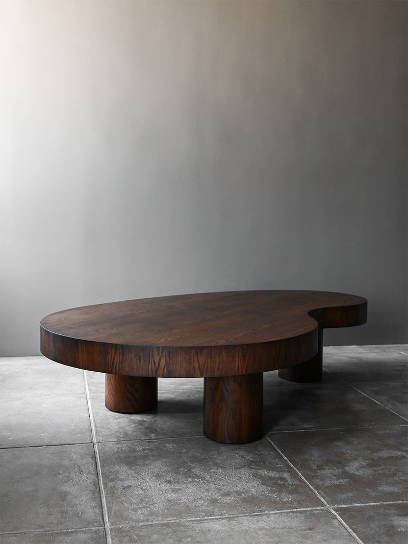 The Norman Stained Oak Coffee Table by Studio OSKLO features a low, mid-century silhouette and an organic, dark-stained top, set on a gray tiled floor against a plain light gray wall.