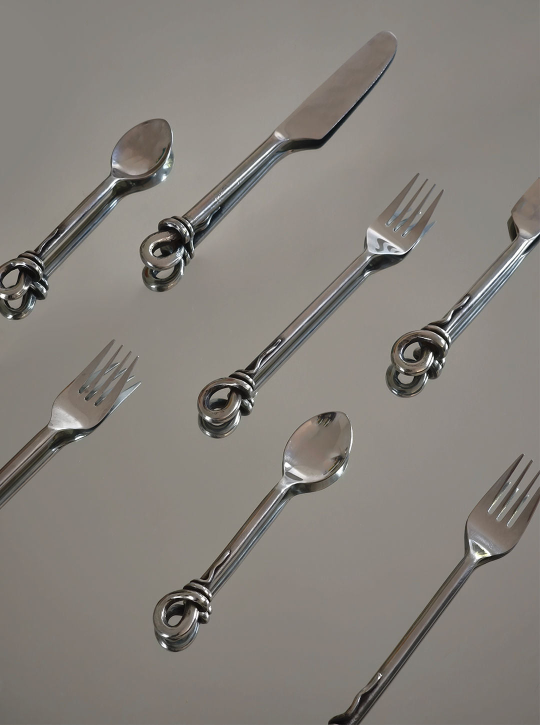 A collection from Les Objoies, the Set of 3 Twist Cutlery features forks, knives, and spoons with ornately twisted handles in an organic design, elegantly displayed diagonally on a smooth, reflective surface.