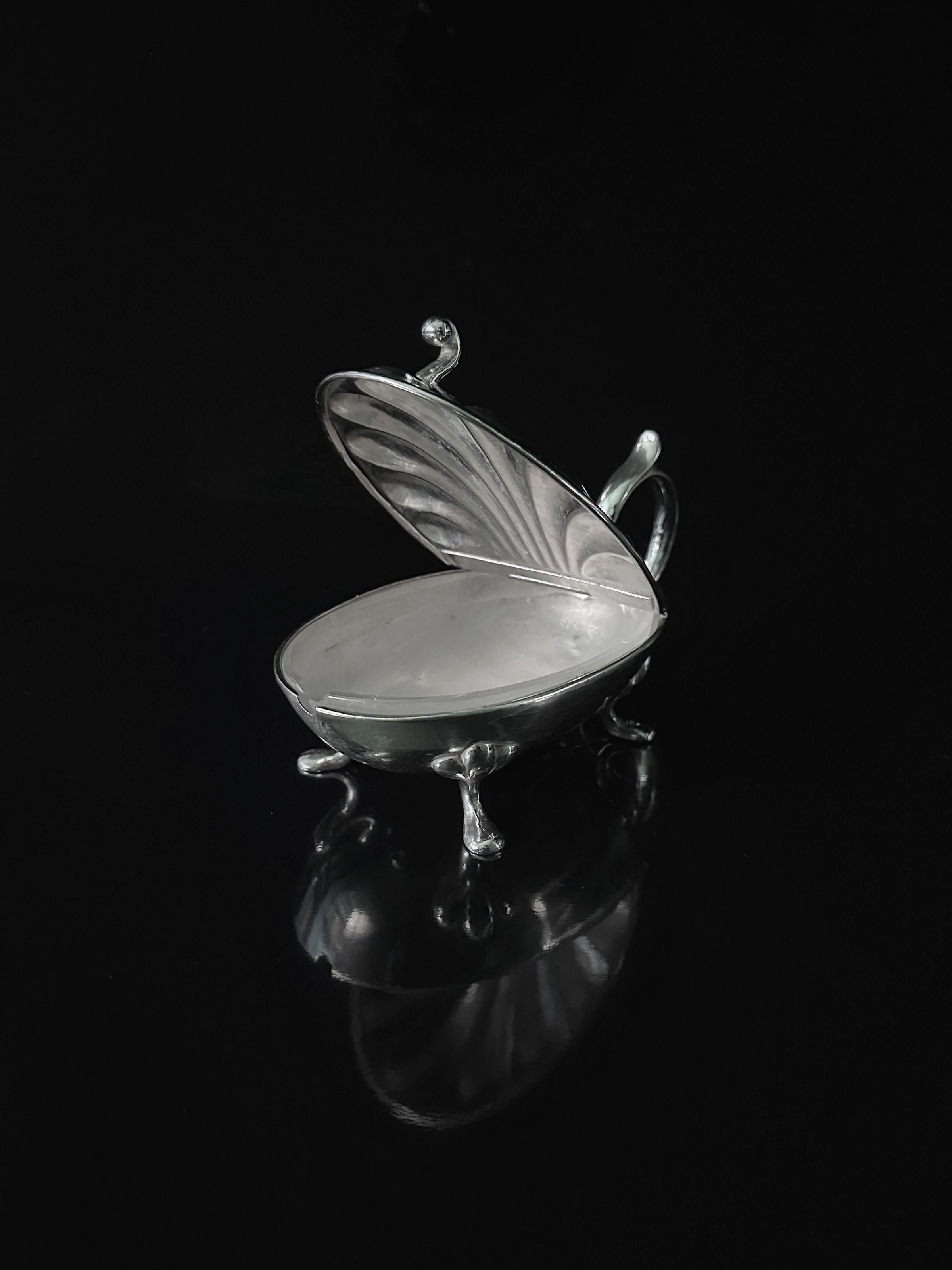 The Dodo Vintage Silver-plated Caviar Frog from the 1950s is an elegant shell-shaped container with fine detailing and four decorative legs. Its hinged lid opens to reveal a smooth interior ideal for delicate treasures or caviar, sitting beautifully on a glossy black surface.