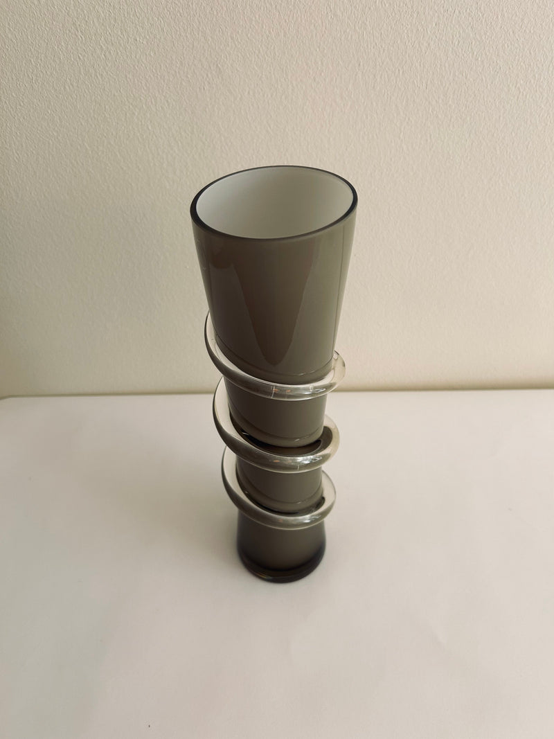 A one-of-a-kind, contemporary vase from Flock Studio, featuring a gray hourglass shape with three horizontal rings of smokey glass spiraling around its center, elegantly displayed against a simple light background.