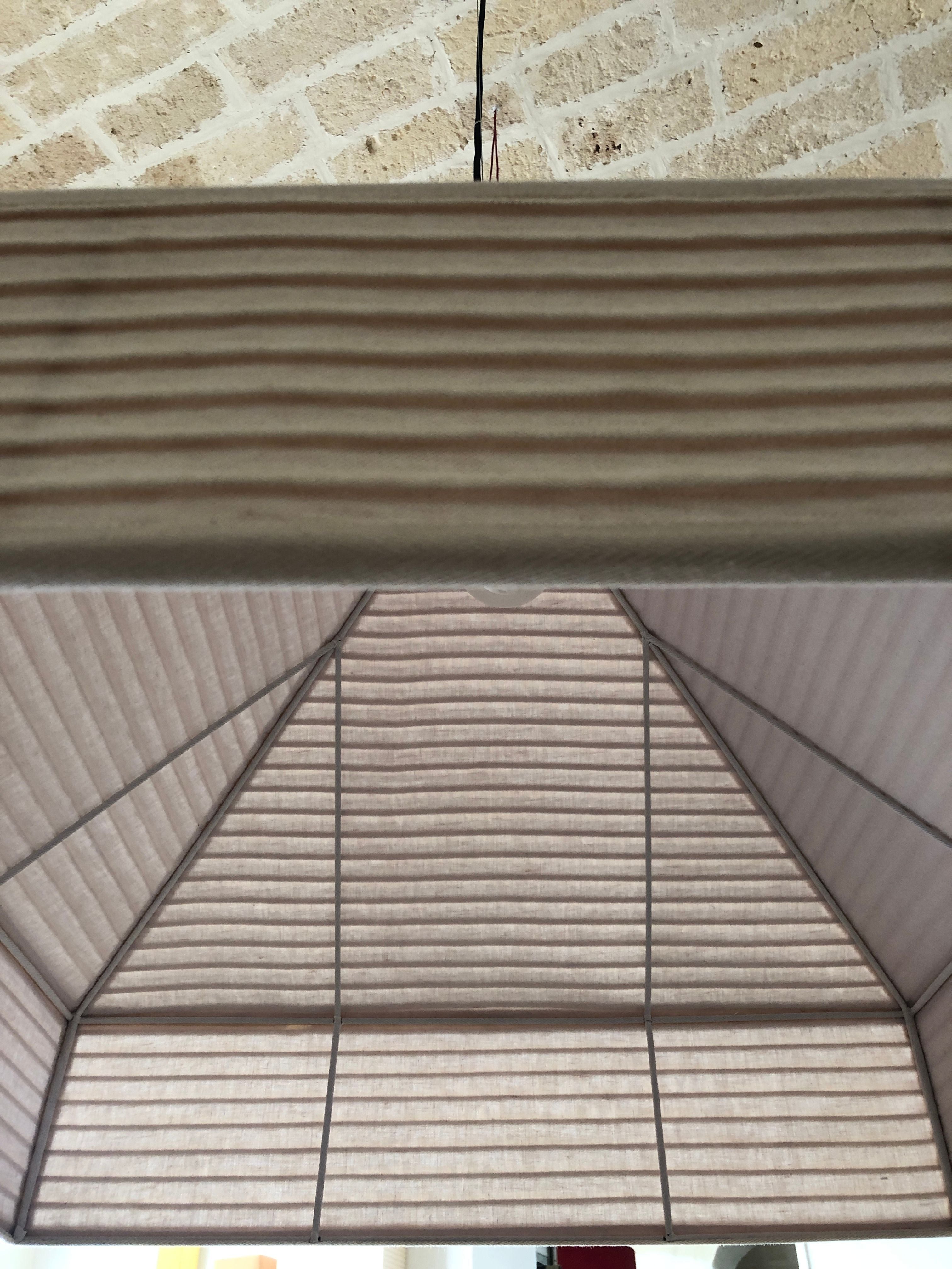 A close-up of a Pyramid Hanging Lamp by Nassi Lamps hanging from a ceiling with a textured surface. The fixture has a geometric, ribbed structure, with a rectangular frame and a pyramid-like shape inside. The light is off, and wiring is visible above it.