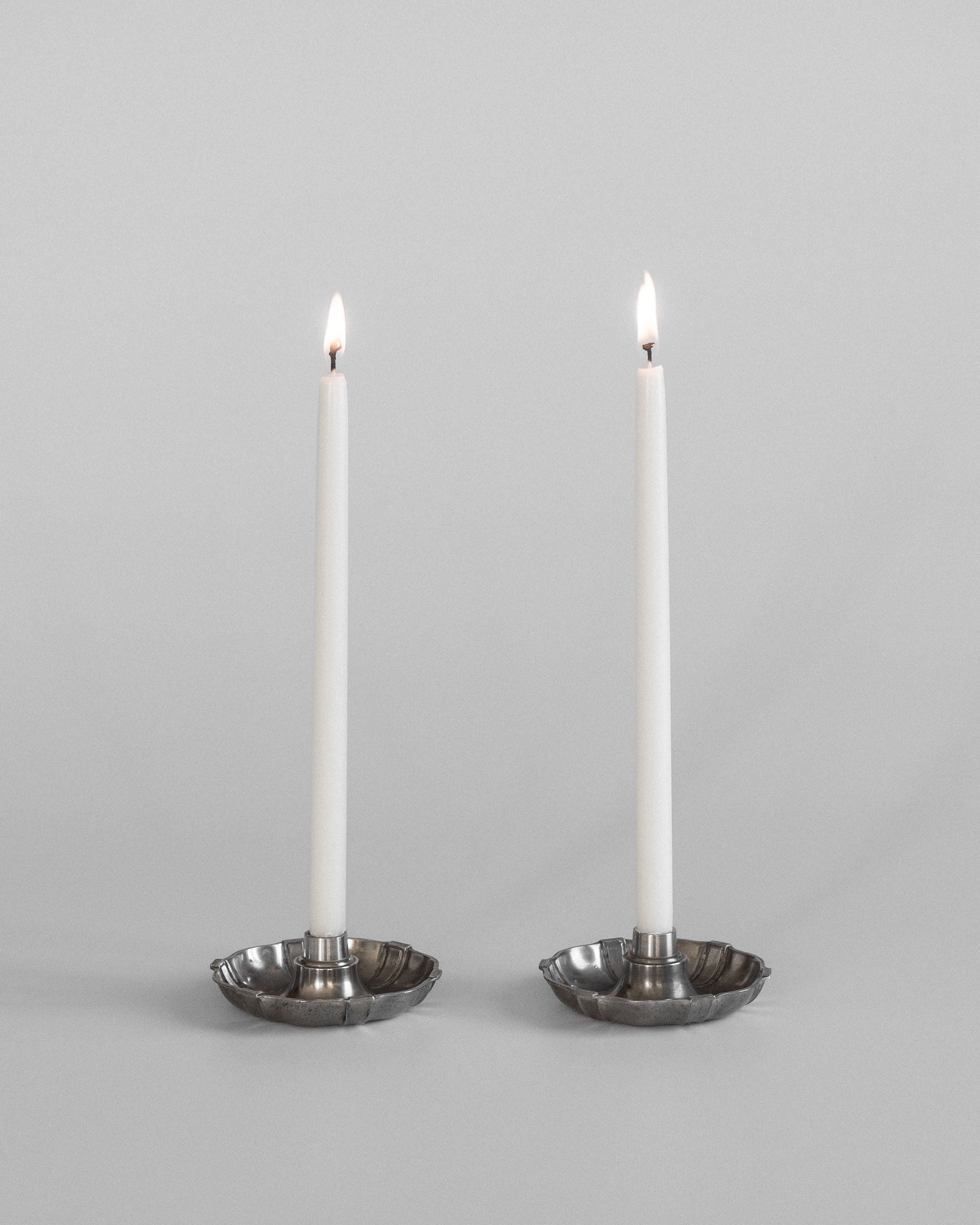 Two lit white candles are elegantly set in Spigel's Modernist Metal Candleholders, 1930s, against a plain, light gray backdrop. The small and steady flame of each candle casts a gentle glow reminiscent of C.G. Hallberg's Swedish Modernist metalwork.