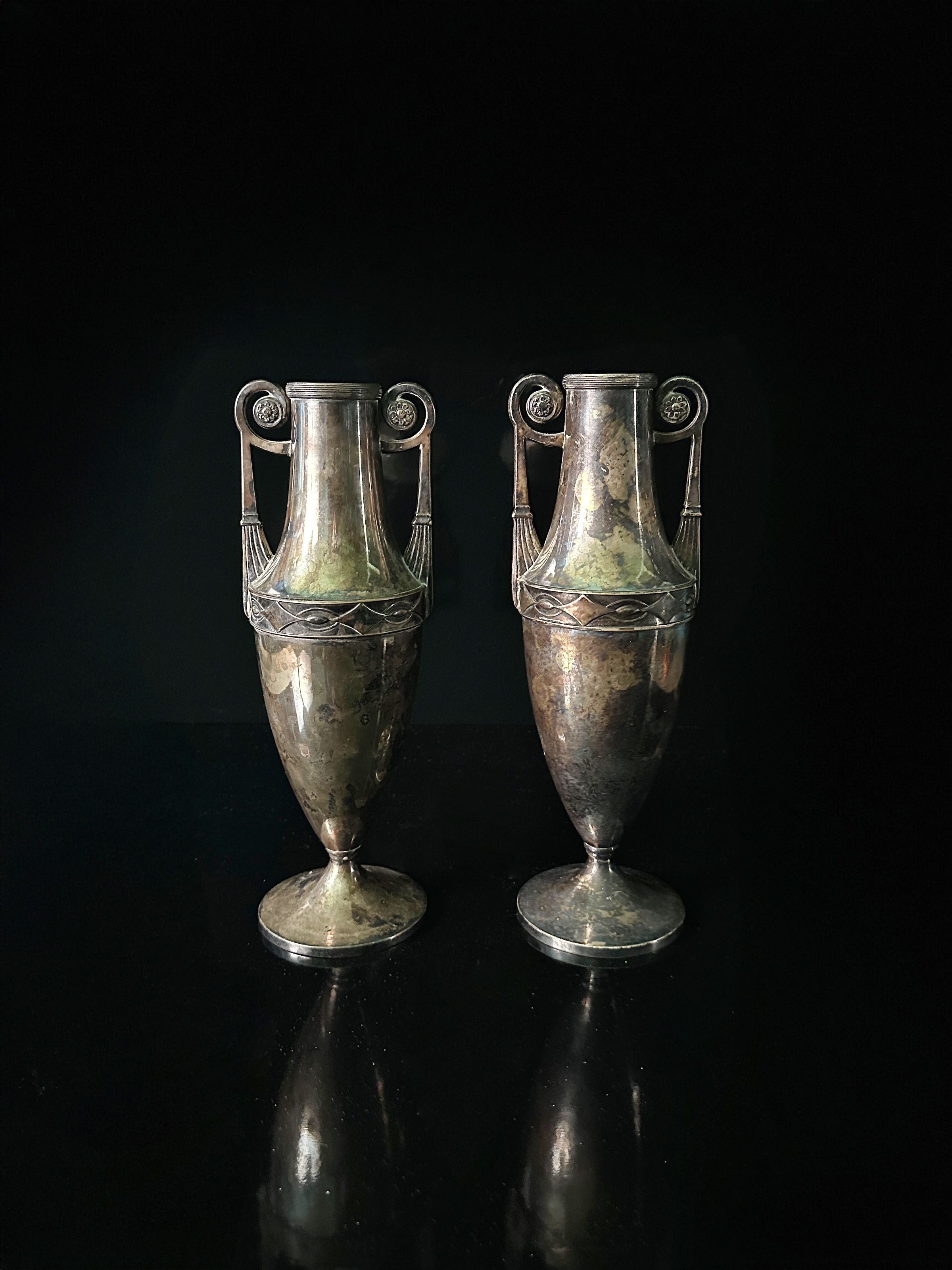 A pair of Dodo Vintage's silver-plated Art Nouveau Amphora from 1890, featuring ornate handles and detailed engravings, stand against a dark background. Their aged patina enhances their vintage charm.