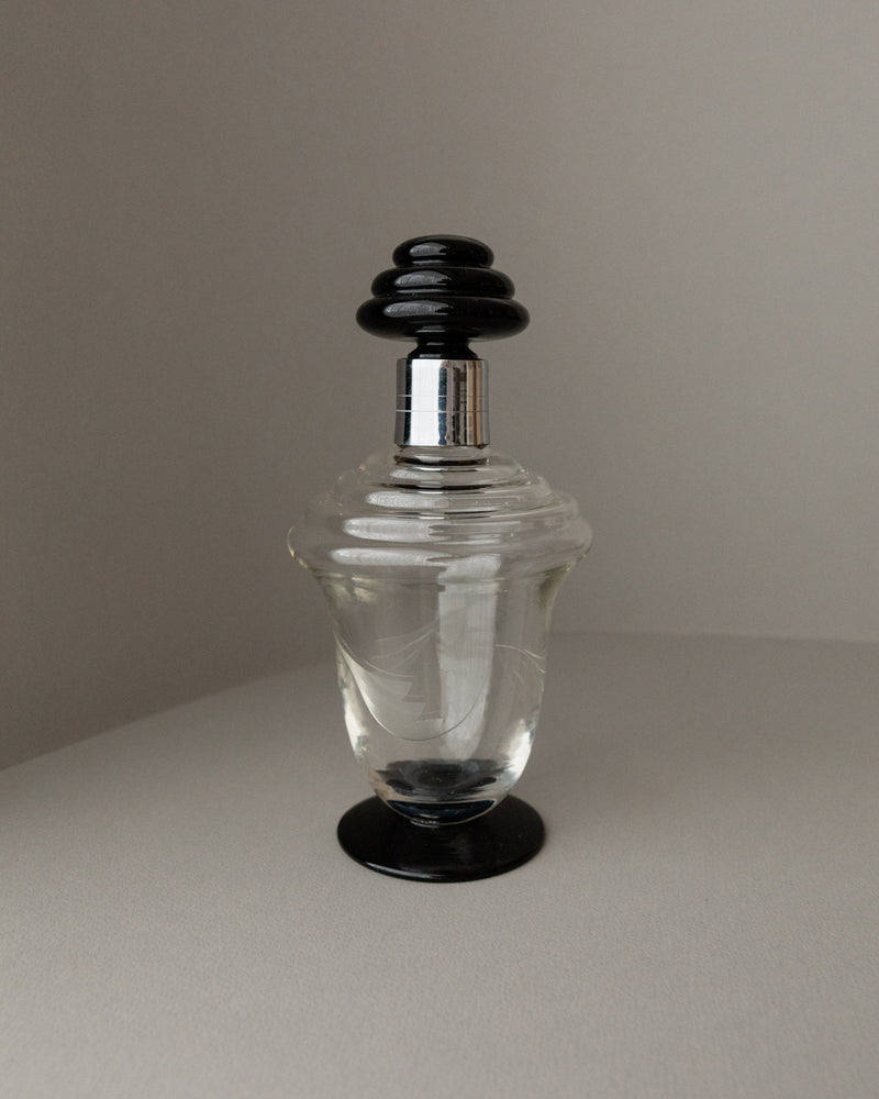 The Murano Glass Decanter by Bottega Jacobs, featuring a wavy pattern and clear body inspired by 1930s designs, is topped with a sleek black cap and elegantly set against a gray background on a neutral surface.