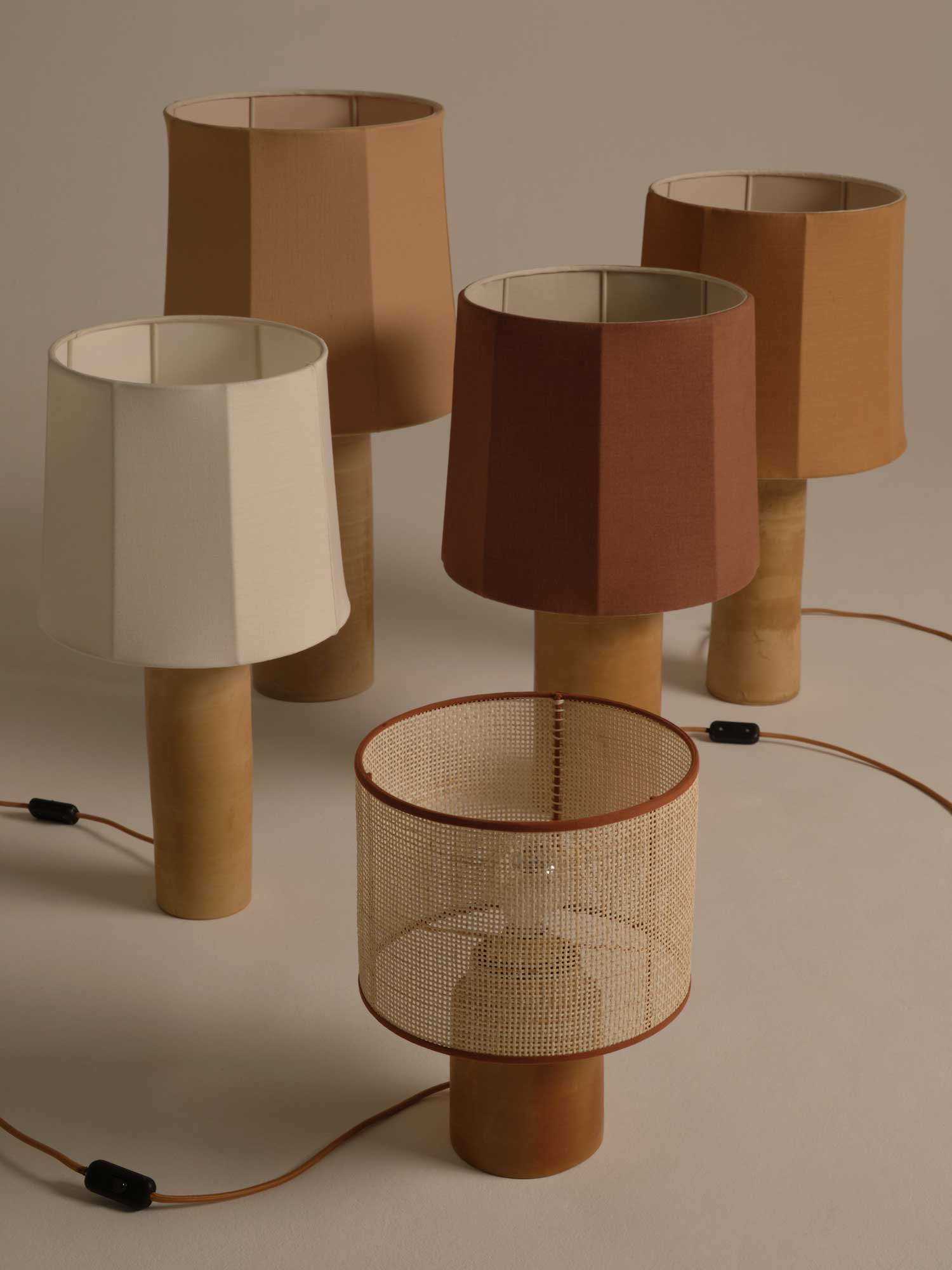 A collection of five Sonora Linen Table Lamps by Nassi Lamps with cylindrical wooden bases is displayed. Four lamps feature fabric shades in varying earthy tones, reminiscent of Puglia's rustic charm, while one lamp boasts a woven rattan shade. Brown electrical cords with black switches are visible, lying on a beige surface.