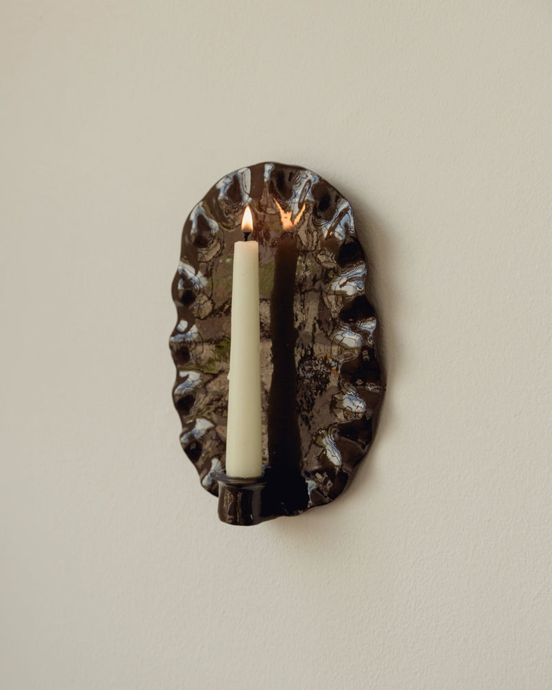 Ortigia oval candle sconce with lit taper candle on beige wall