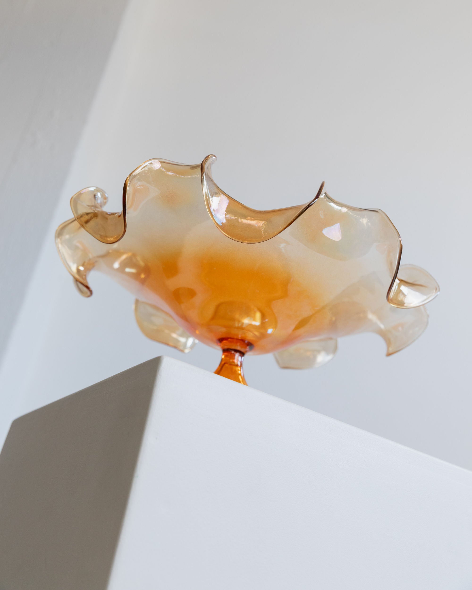 A delicate, translucent decorative piece with wavy, ruffled edges sits atop a white display pedestal. This vintage Murano Glass Bowl by Parise Vetro 70s from Bottega Jacobs boasts a warm amber hue, catching the light to create an orange iridescence against the smooth, light-colored wall background.