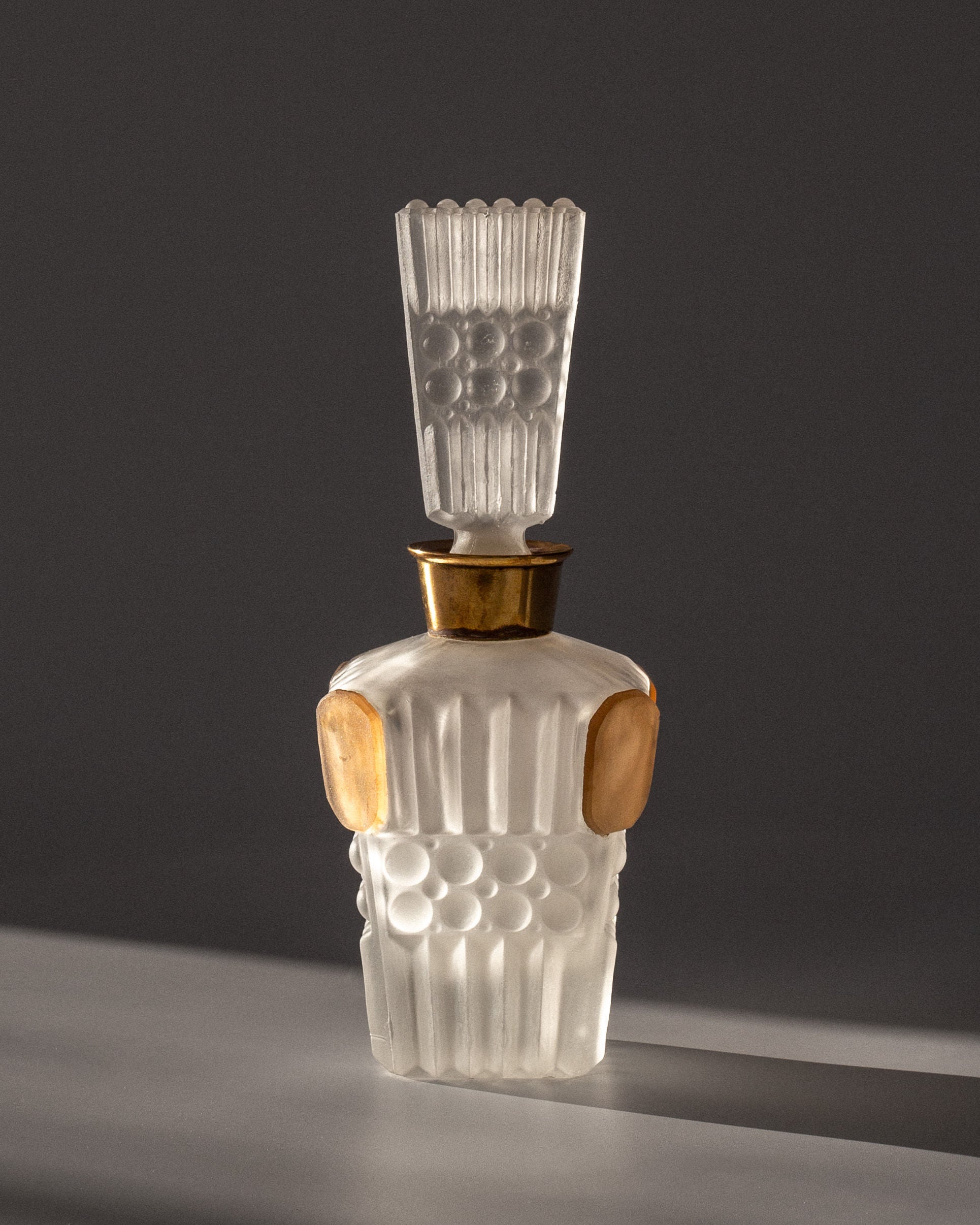 The Spigel Art Deco Perfume Bottle, crafted in 1930s France from frosted glass, features an ornate elongated stopper and raised circular geometric patterns with gold accents. It sits elegantly against a dark gray backdrop, emphasizing its timeless elegance.