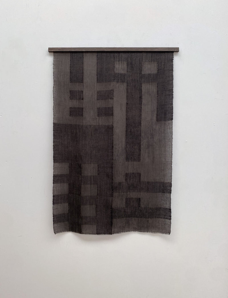 The Grid I tapestry by Sundance Studio, inspired by the Bauhaus movement, hangs on a wall. It features geometric rectangles and lines in textured gray for a modern, minimalist look.