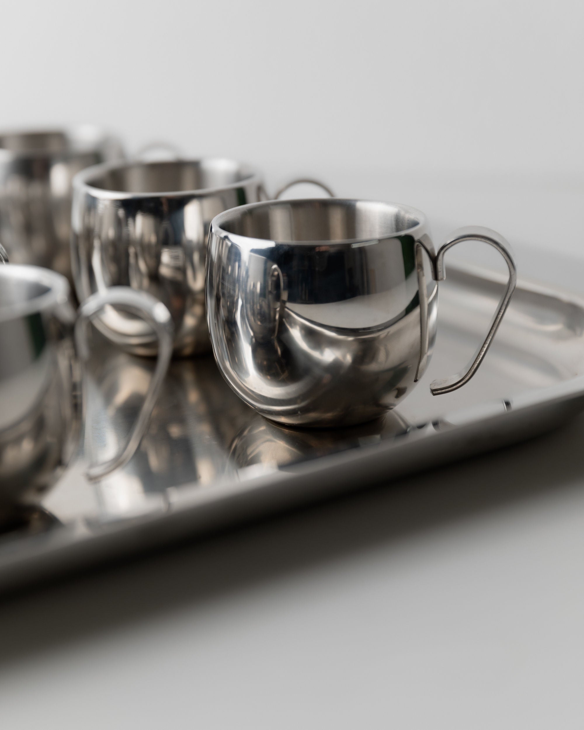 A close-up of the Bottega Jacobs Coffee set by IMCO 70s, featuring polished stainless steel teacups with handles on a matching metal tray. The reflective surface enhances its modern, minimalist design for a sleek look.