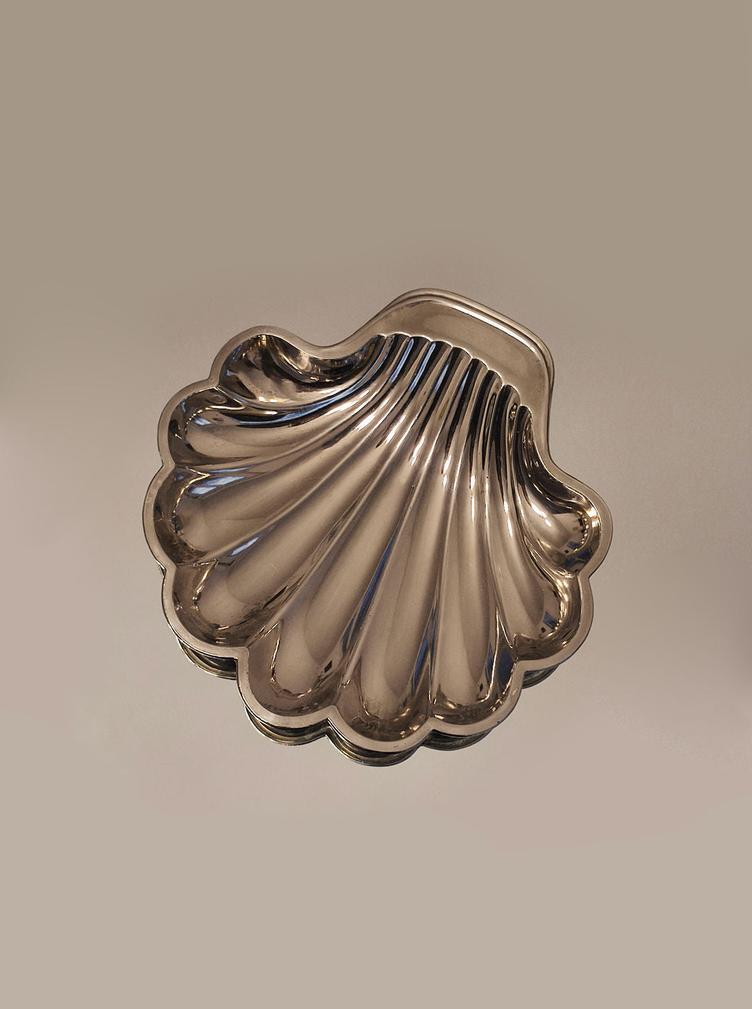 The silver-plated Shell Shape Vide-poches by Les Objoies glistens with its shiny surface, detailed ridges, and smooth handle, exuding elegance against a plain beige background.