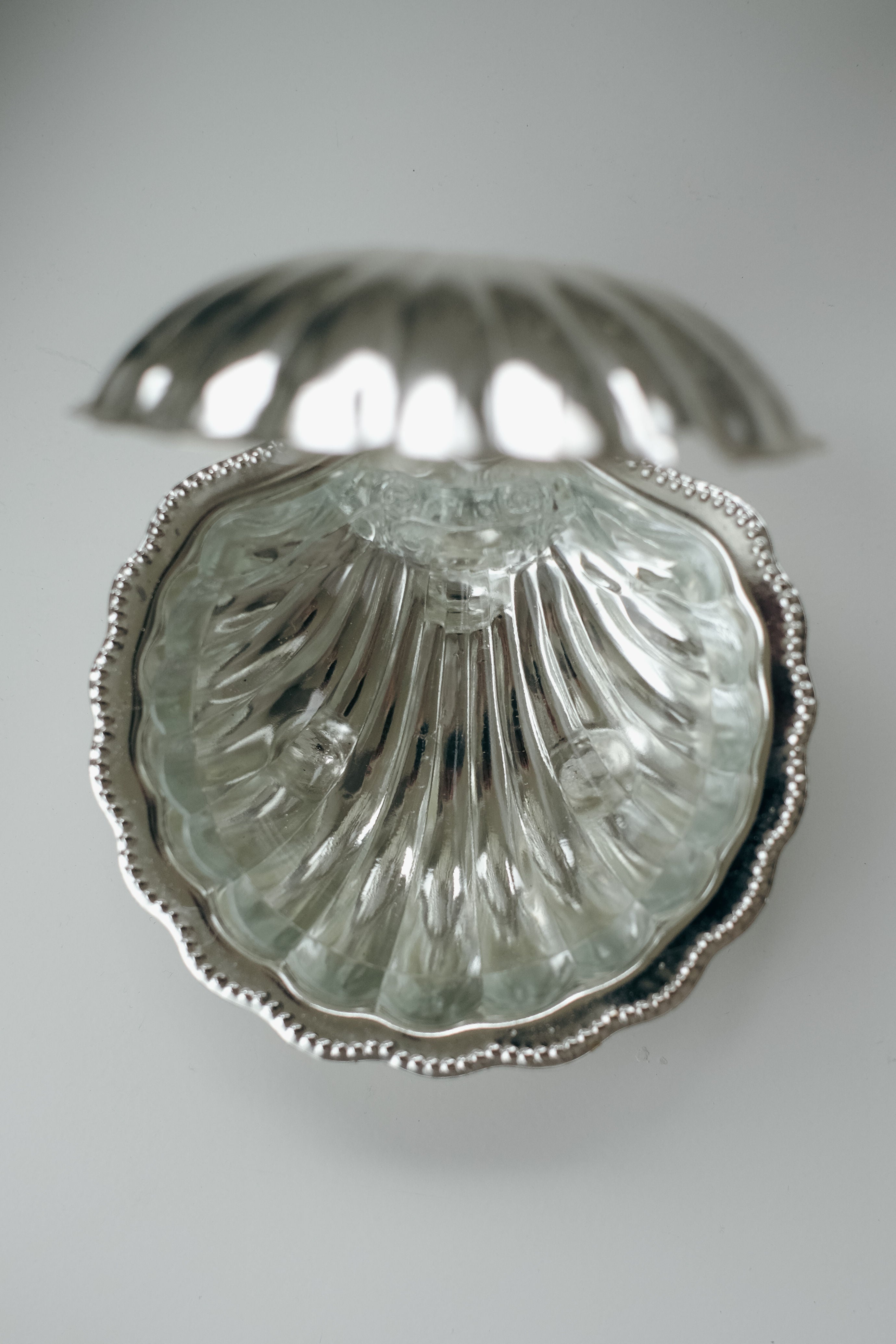 The open Shell Shaped Butter Dish by septembre studios, with its metallic seashell design and textured, reflective interior with a beaded edge, shines like a vintage brass jewelry box. The lid tilts back slightly to reveal the empty shell-like compartment against a softly lit backdrop.