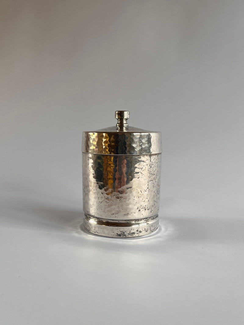 A small, cylindrical Art Deco Hammered Pewter Box by Gero from Maud Vaughan, featuring a lid with a knob, sits against a plain light-colored background, exuding elegance.
