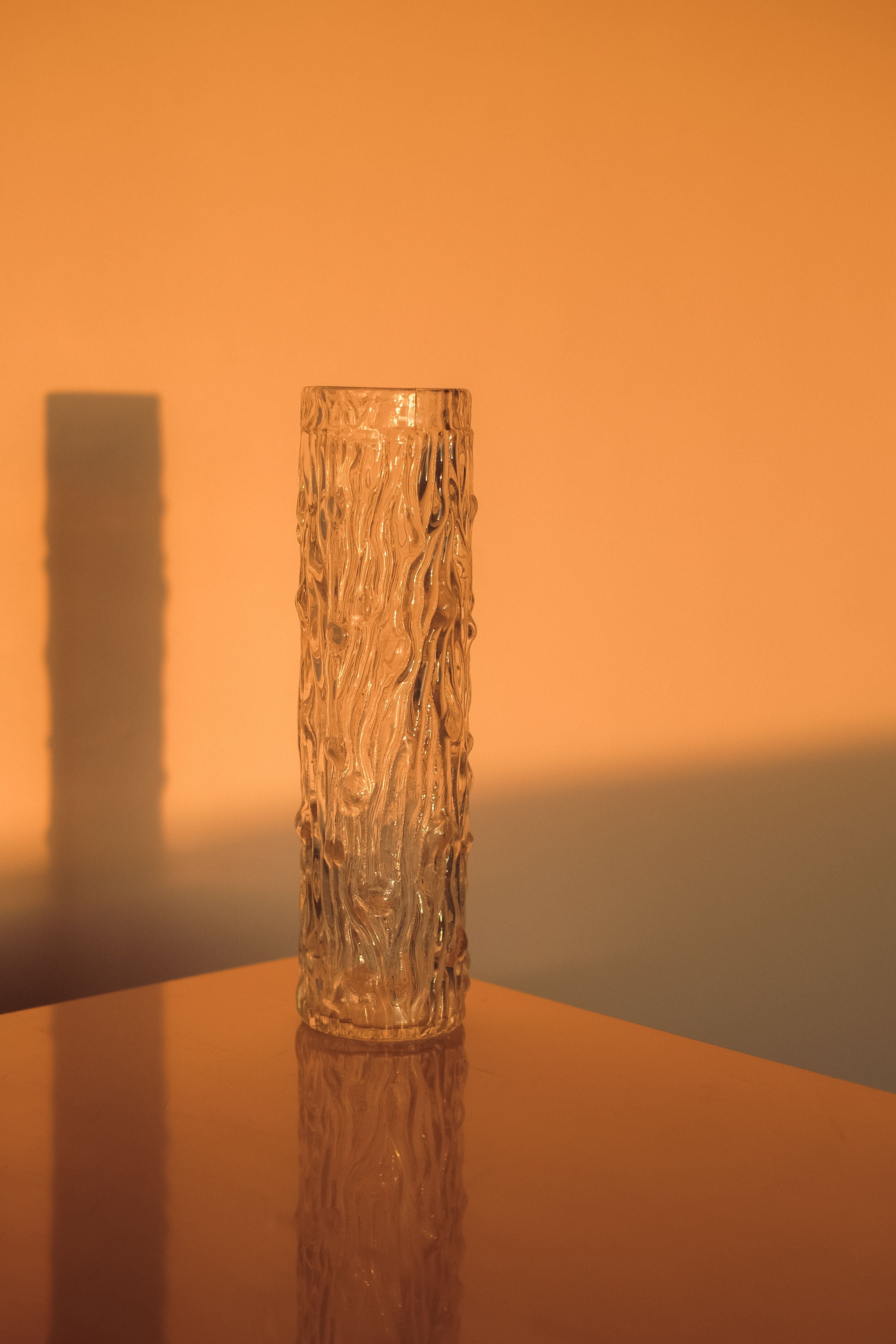 A small septembre studios glass cylinder vase with a bark-like wavy pattern stands on a reflective surface, bathed in warm, golden sunlight that casts a distinct shadow against the orange-toned backdrop.