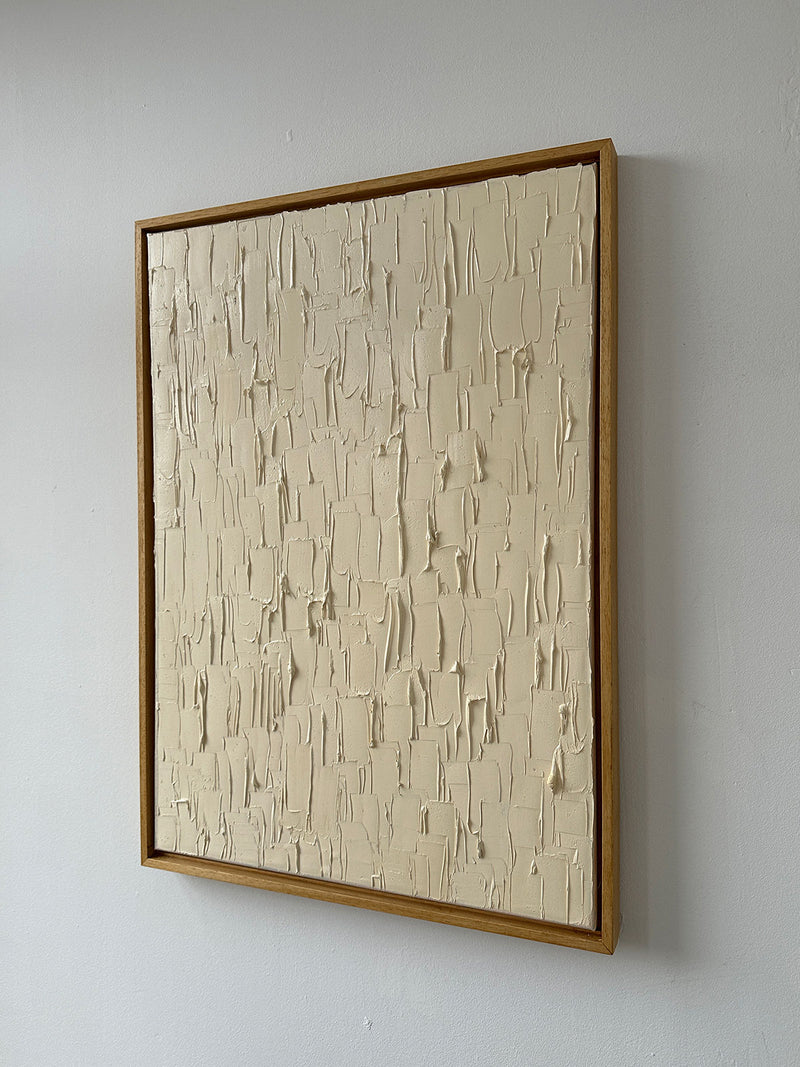 The "Warm White" oil on canvas by Roi Elmaliah showcases a textured cream-colored surface with irregular, raised patterns. This captivating piece is displayed against a plain white wall, evoking the minimalist aesthetic of an Amsterdam gallery.