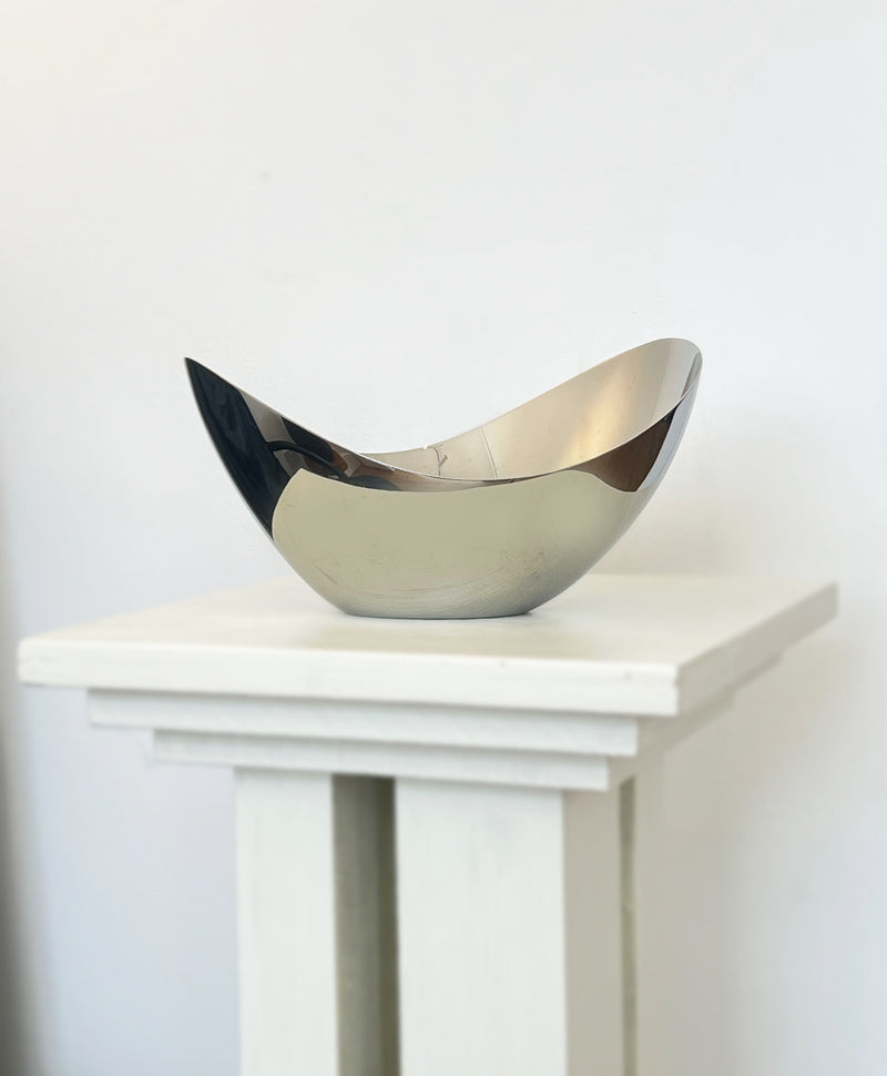A shiny, metallic Porter Studio George Jensen Bloom Bowl sits asymmetrically on a white pedestal, its stainless steel surface reflecting light and surroundings for a modern, timeless, and minimalist look.