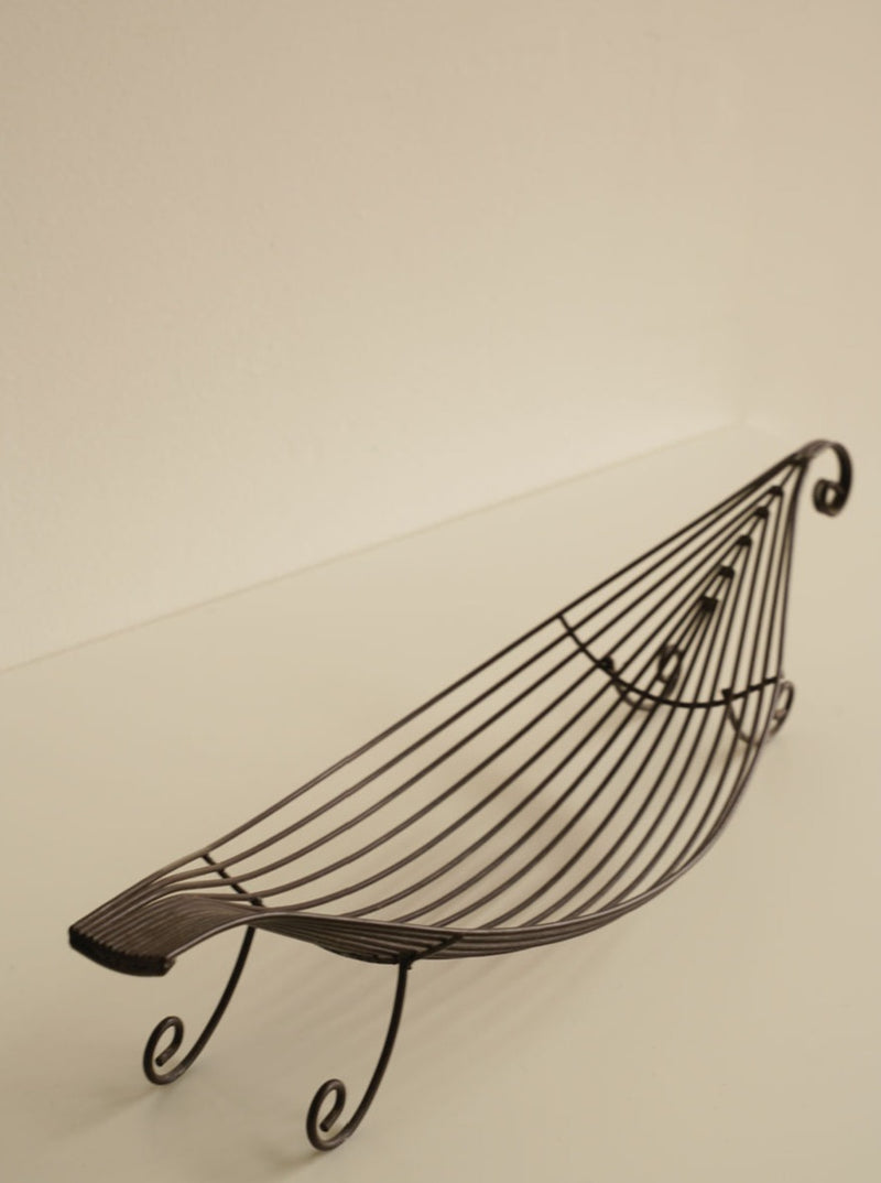 An Iron Swirl Fruit Bowl by Les Objets Amsterdam, designed to resemble a boat with curved lines and a lattice pattern, is placed on a light-colored surface. The elegant fruit bowl has raised ends and sits on small spiral-shaped legs, giving it added charm with its swirl accents.
