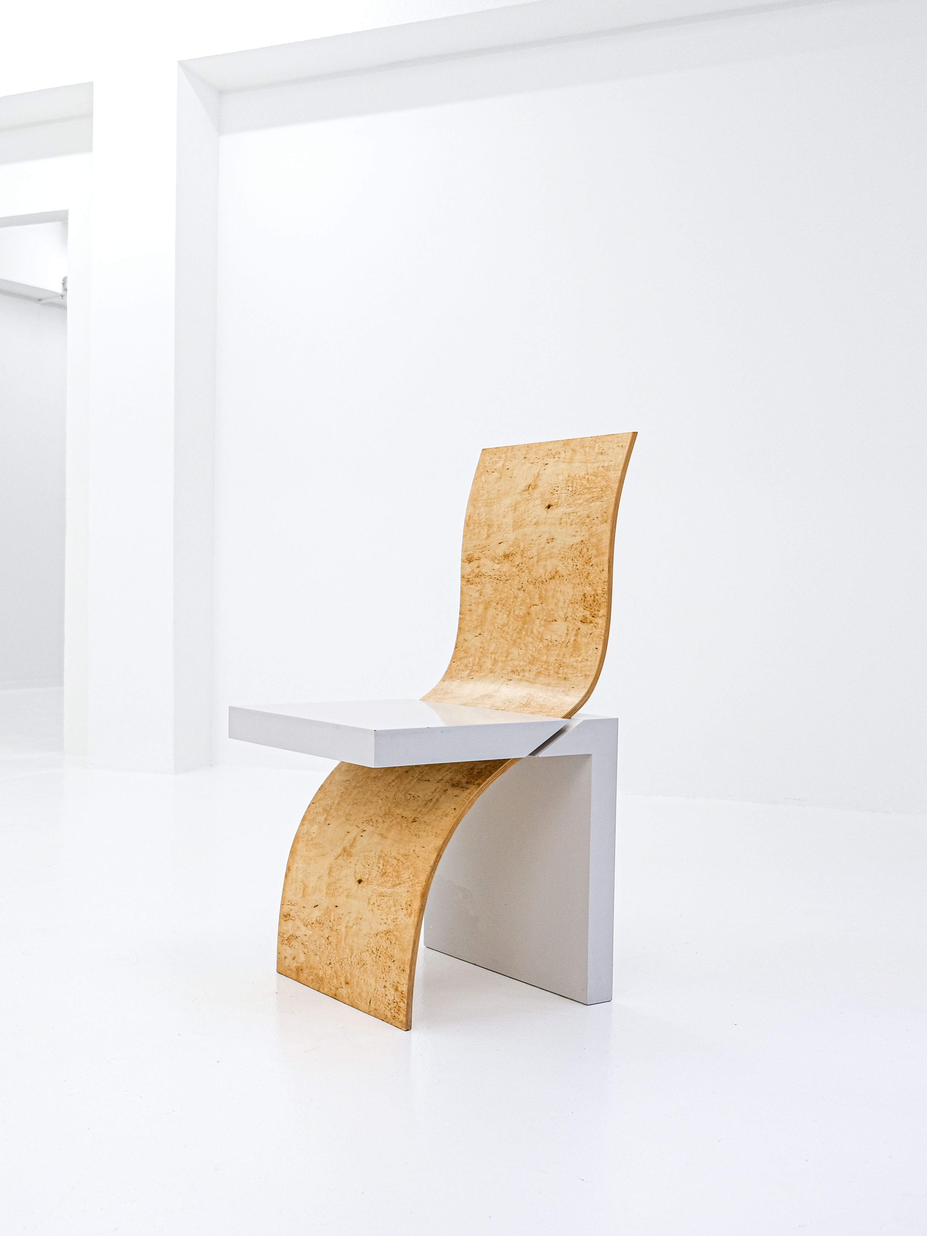 The Postmodern Leda Chair by Angela Oedekoven for antibeige, made in 1990, features a minimalist white rectangular frame with a curved wooden seat. Its seamless transition from seat to backrest stands out beautifully in a stark white room.