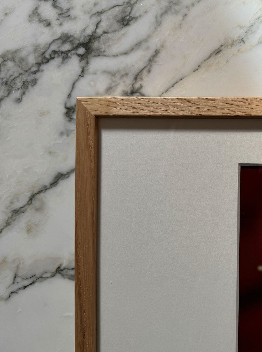 Close-up of a wooden picture frame corner resting on a marble surface. The fine art print inside the frame reveals part of **Orange and Pearls by Adeline Mai**, bordered by a white mat. The frame has a smooth, natural wood finish. The marble background features grey veining and enhances the artwork's elegance.