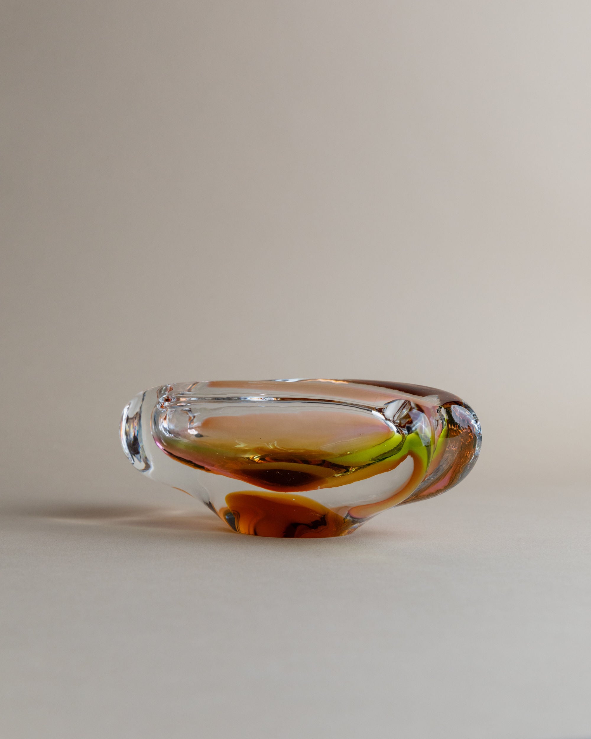 A Glass ashtray by Josef Rozinek for Novy Bor, from Bottega Jacobs, sits on a light beige surface. The smooth, clear glass with swirling colors of amber, green, and red creates a fluid, vintage design against the plain neutral background.