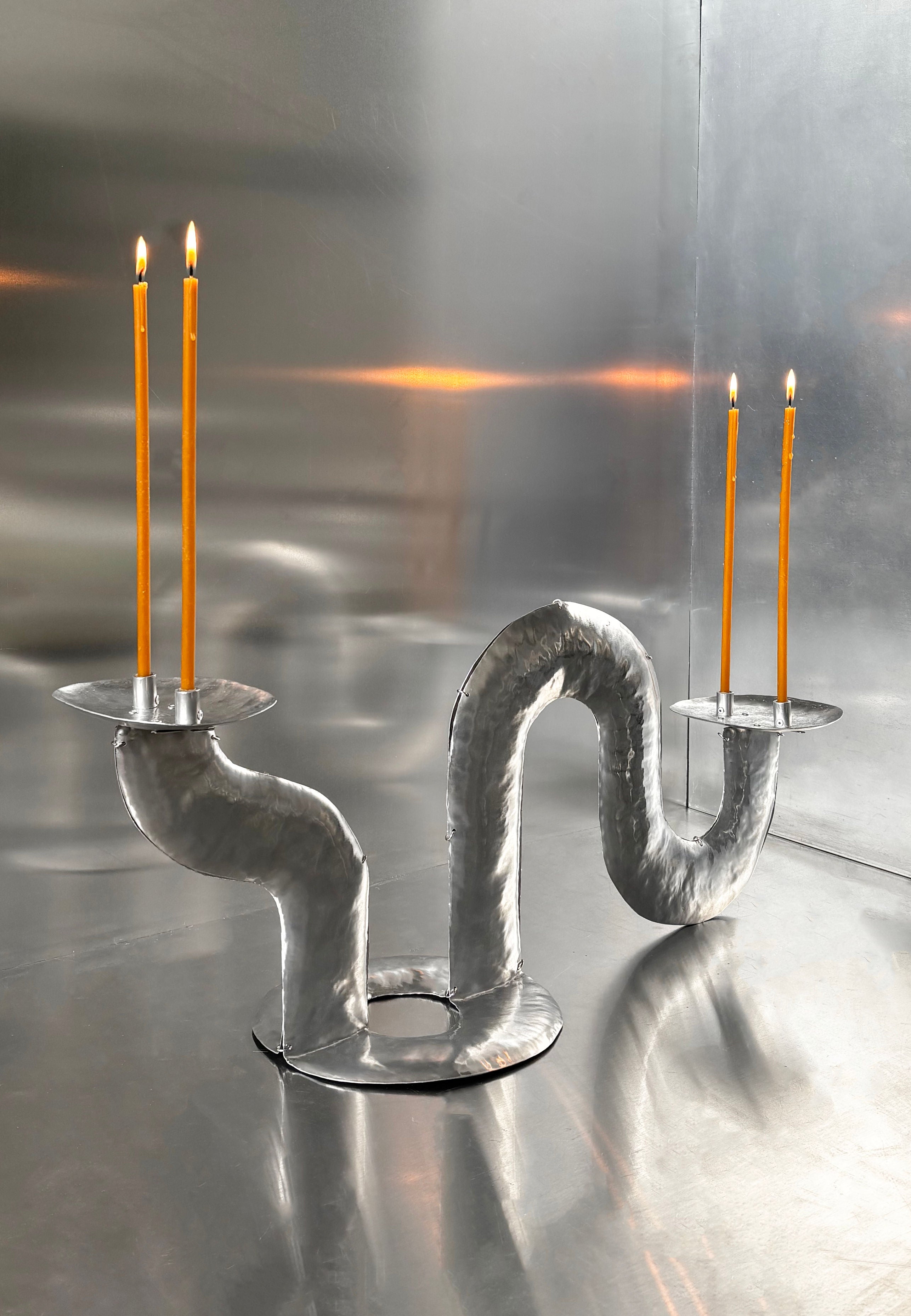 The Phaedra candle holder TABLE by Bregje Sliepenbeek, handmade in Amsterdam, has a twisting metallic design that elegantly holds four slender orange beeswax candles. Its abstract, wavy form creates a captivating silhouette against the sleek, reflective backdrop.