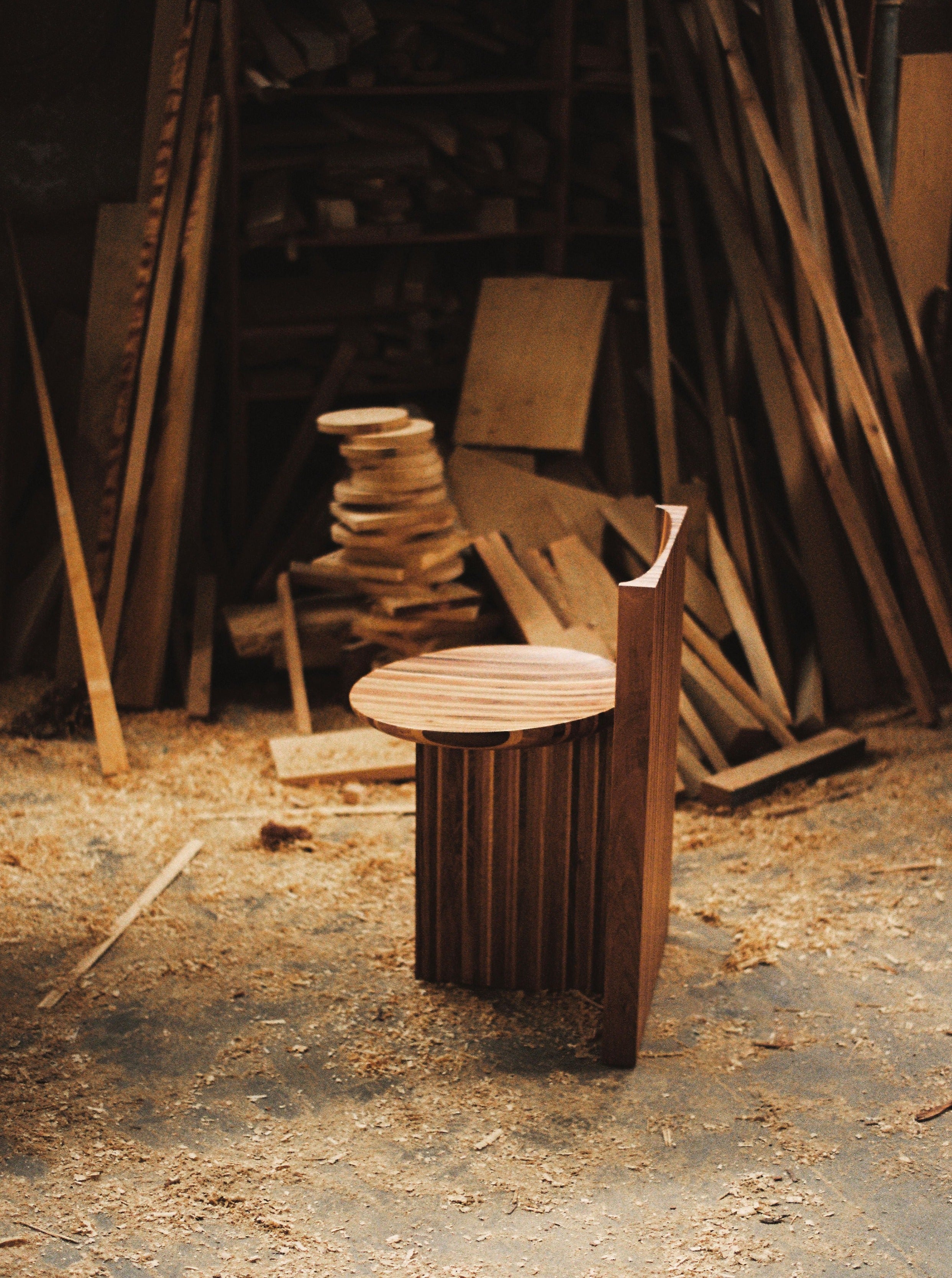 The Vaga Chair by Rosana Sousa stands alone in a workshop filled with wood planks and sawdust. Handcrafted from solid walnut and oak, the chair features a half-circle backrest and ribbed base, showcasing its modern aesthetic. Various woodworking tools and materials are scattered in the background.