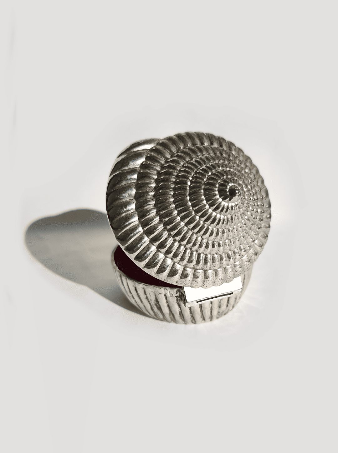 The Silver-plated Shell Box by Les Objoies, featuring a circular spiral seashell design, casts a shadow on a plain white surface.