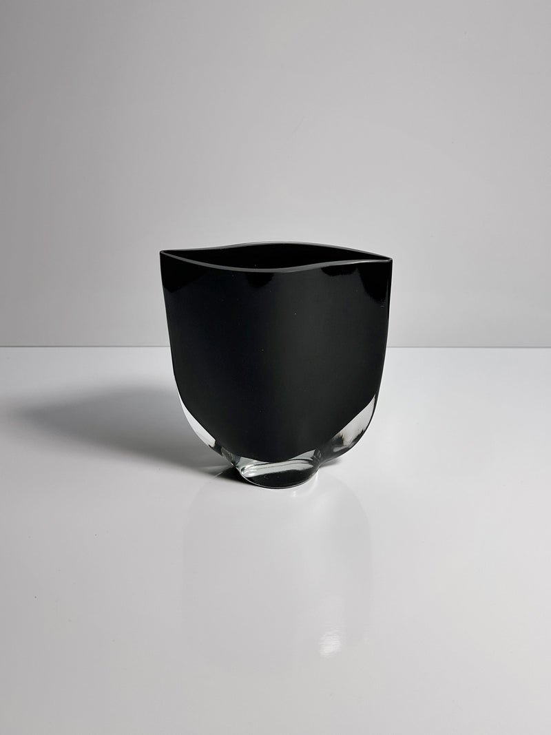 A Black Glass Vase by Kaj Franck from Dodo Vintage, with a glossy, irregularly shaped form and a clear, slightly textured base, sits on a white surface against a plain gray background. This vase embodies modern minimalist aesthetics reminiscent of Kaj Franck's iconic designs for Nuutajärvi Notjö.