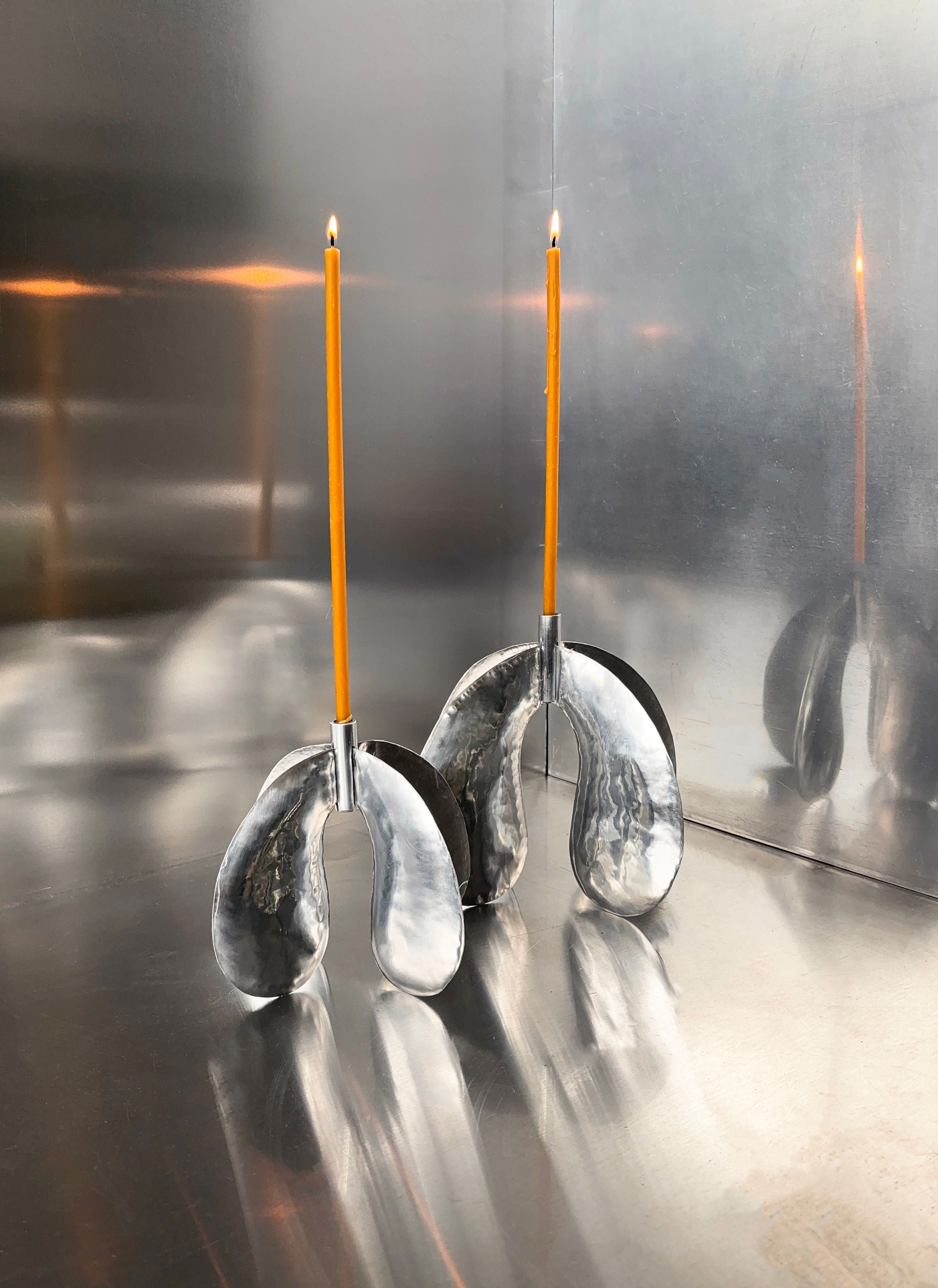 The Phaedra Candle Holder Small by Bregje Sliepenbeek is a modern metal piece, hand-made in Amsterdam, featuring two curved, mirrored bases that support incense sticks. Its reflective surface enhances the artistic effect of wafting smoke, just like a small candle holder's charm.