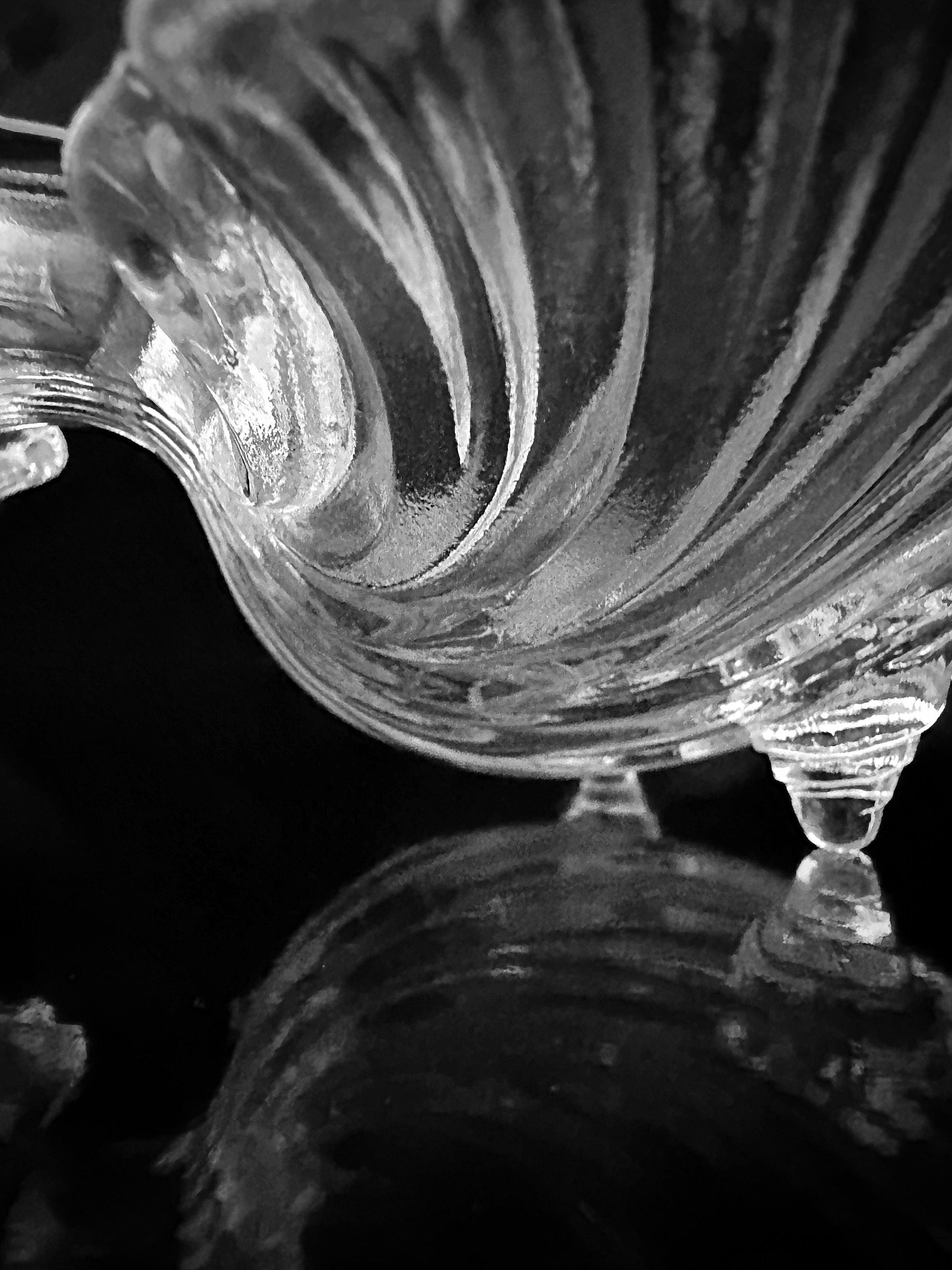 Presenting a close-up black and white image of an intricately designed glass piece from Dodo Vintage, this Shell Shaped Glass Bowl from the 1980s showcases its swirling pattern and reflective surface. The dark background enhances the texture and form, adding a touch of elegance to this delicate presentation.