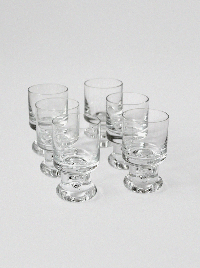 The Tapio Wirkkala Set of Glasses by Vintage Stories features six clear, empty glass tumblers with thick bases in a loose cluster on a plain white background, evoking classic Mid-century style.