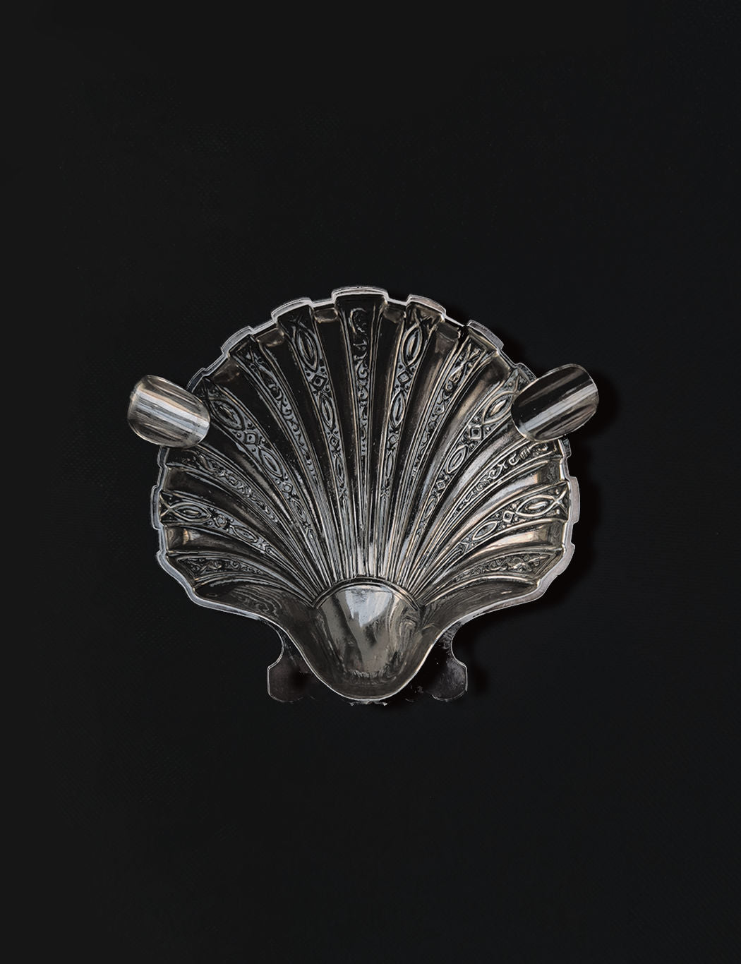 The Shell Ashtray by Les Objoies is an ornate, fan-shaped silver dish resembling a shell, featuring intricate patterns and two small, textured handles on either side, set against a dark background.