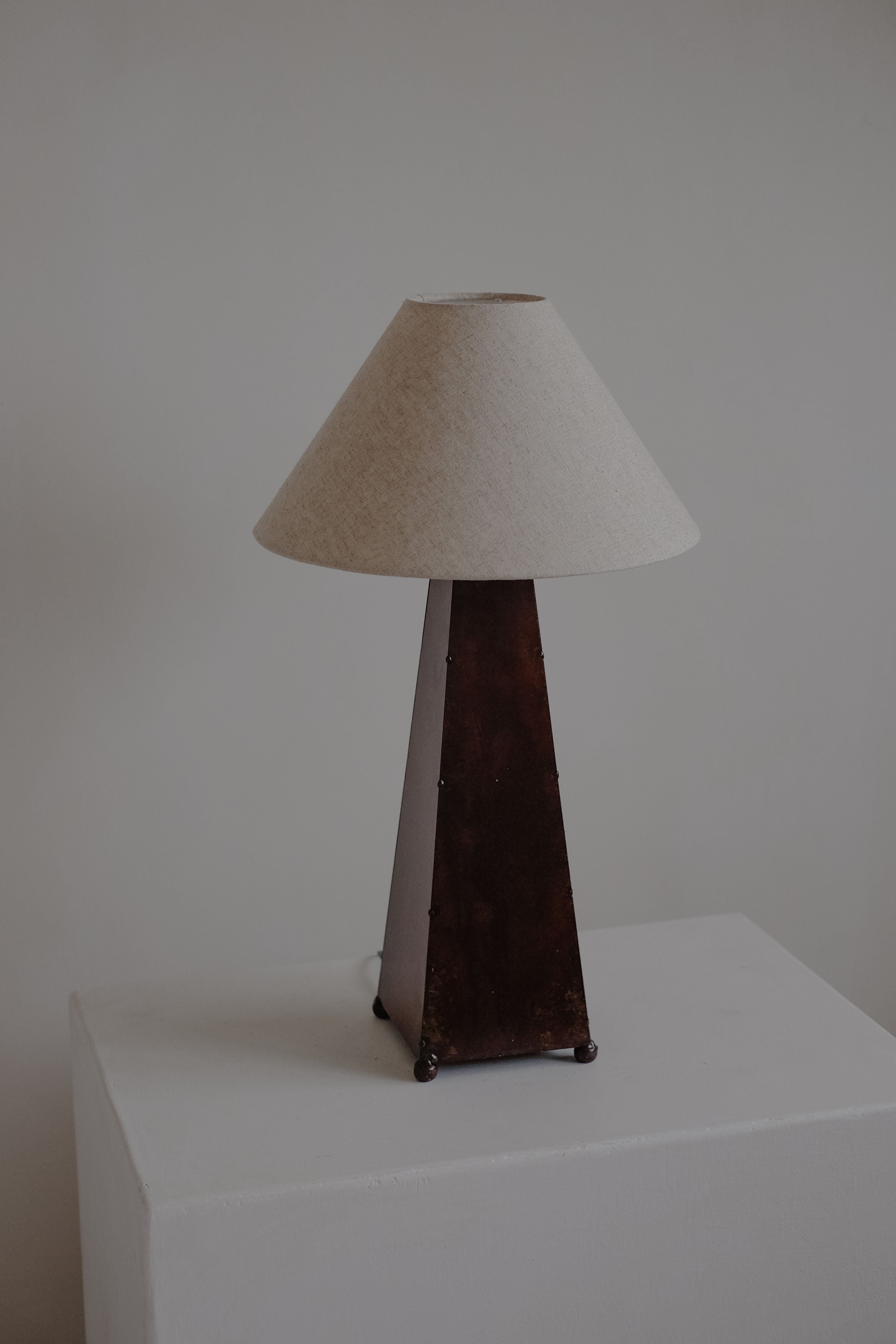 A minimalist Obelisk Table Lamp by septembre studios has a brown triangular base with rust-copper patina and a beige cone lampshade. It sits on a white pedestal against a light backdrop, offering elegance with a dimmable switch.