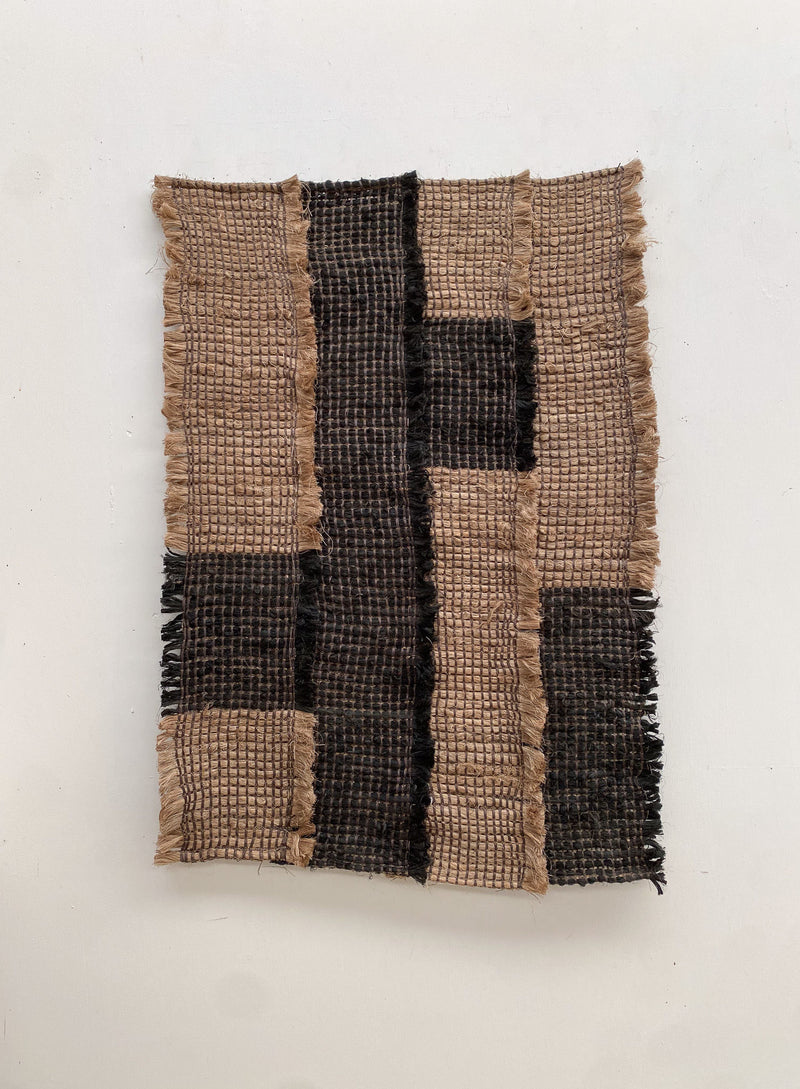 The Ink / Sand from Sundance Studio is a woven textile crafted from banana fiber, featuring a grid pattern with alternating light and dark brown sections. Its frayed edges enhance its rustic texture and highlight its biodegradable qualities.